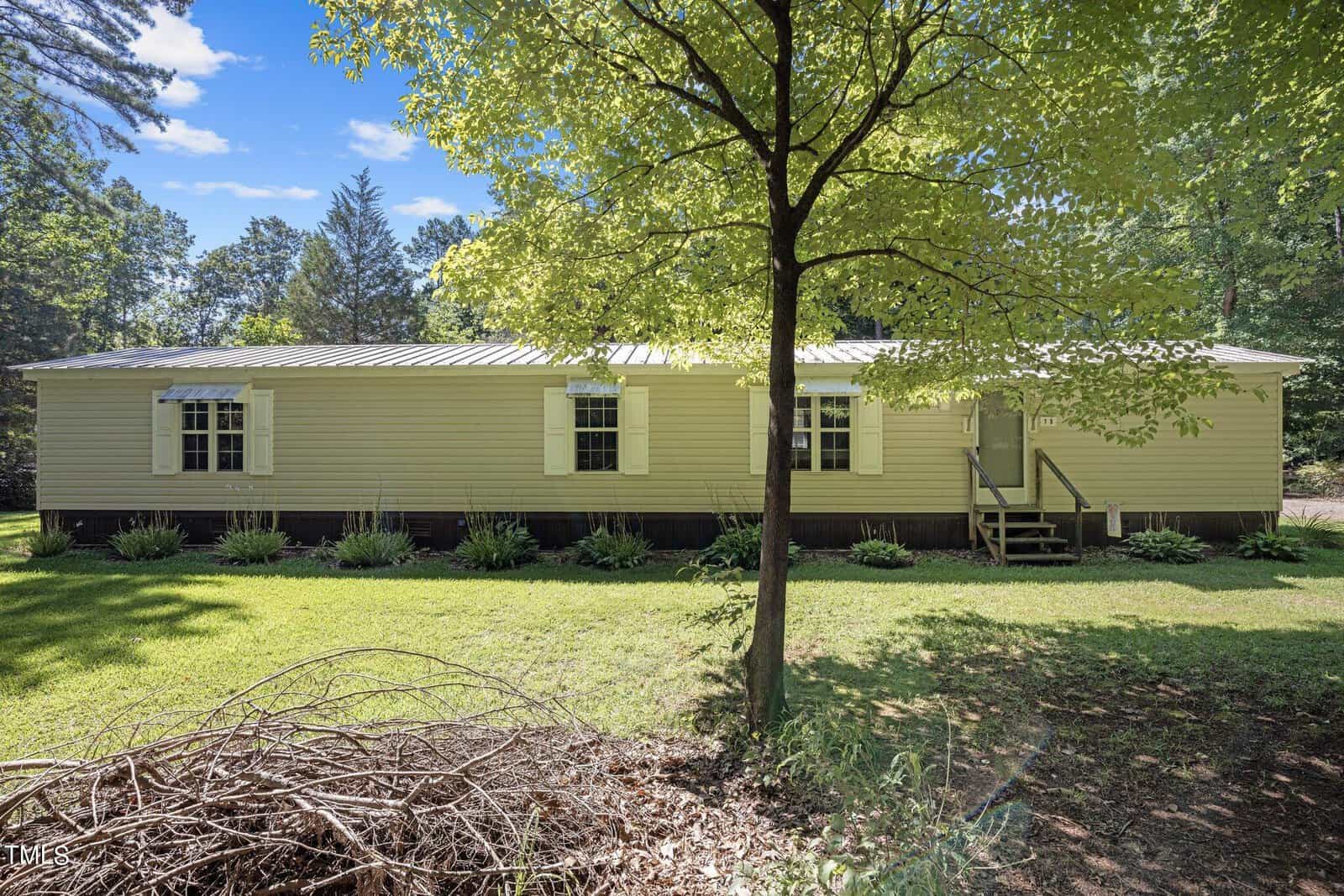 Single-story home with white siding and a metal roof, surrounded by lush greenery and trees. The house features several windows with shutters and a small front porch with steps. The yard is spacious with a pile of branches in the foreground. Bright, sunny day.