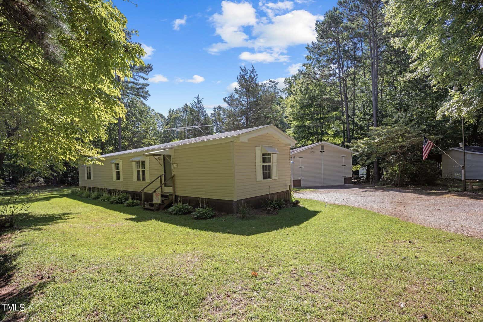 A yellow, single-story mobile home with a small porch and steps, surrounded by lush green grass and trees. A gravel driveway leads to a beige shed in the background. The sky is bright and blue with a few clouds. An American flag is visible near the shed.