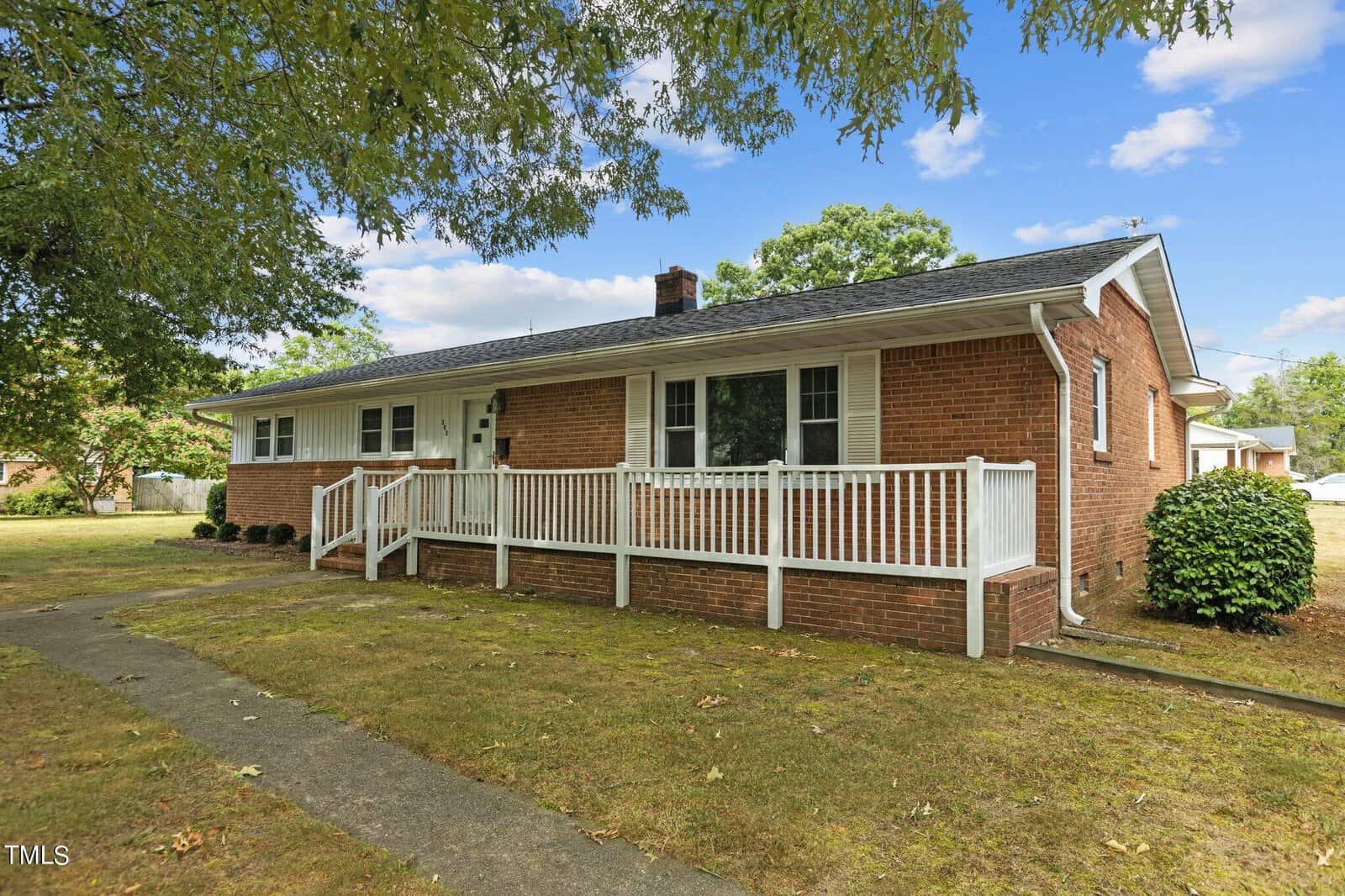A one-story red brick house with a dark shingle roof, white trim, and a long front porch with white railings, surrounded by a grassy yard and shaded by trees.