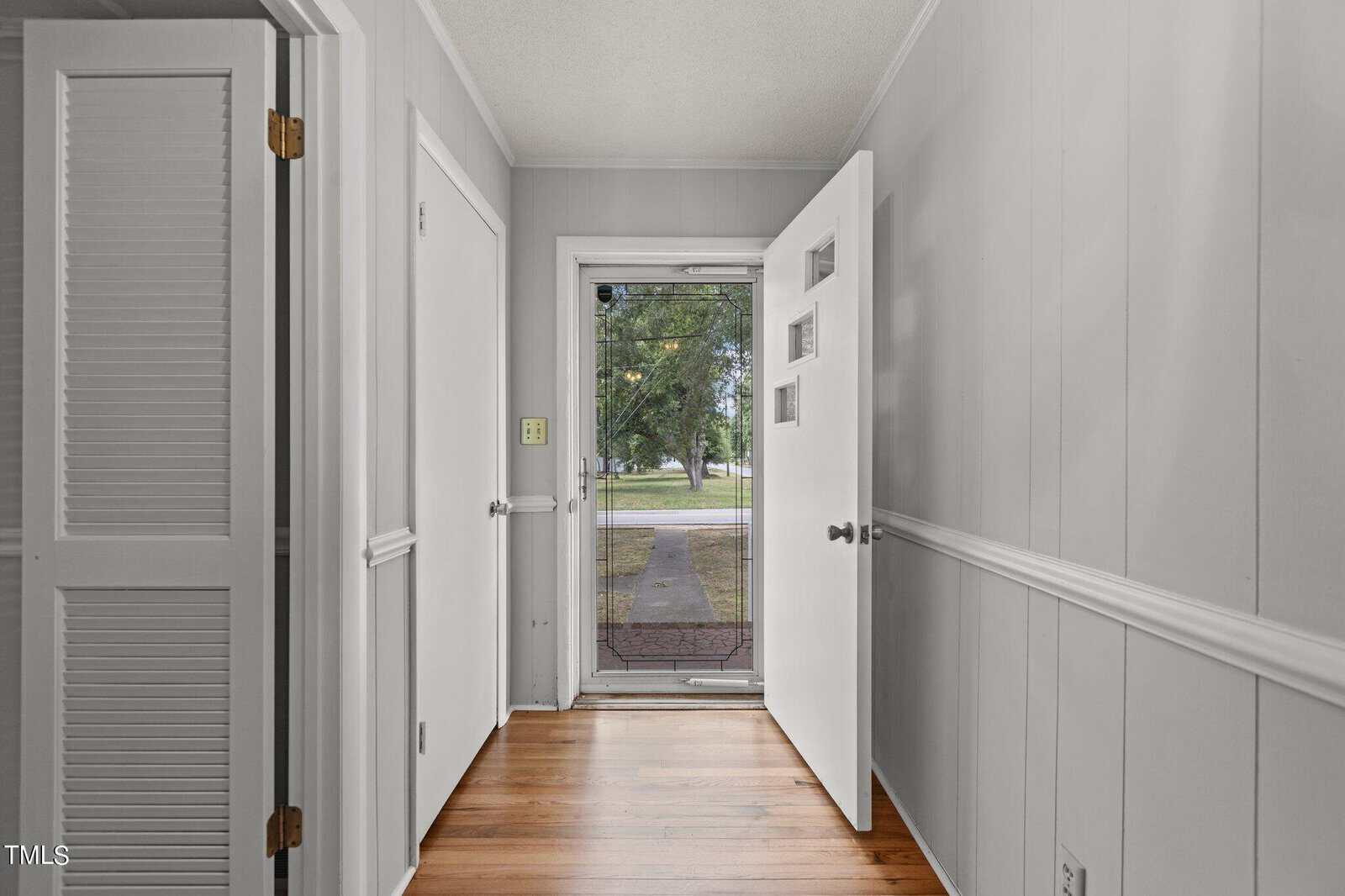 A view of a home's entryway with wood flooring, light-colored paneled walls, and an open white front door. A glass storm door reveals a sidewalk, green lawn, and trees outside.