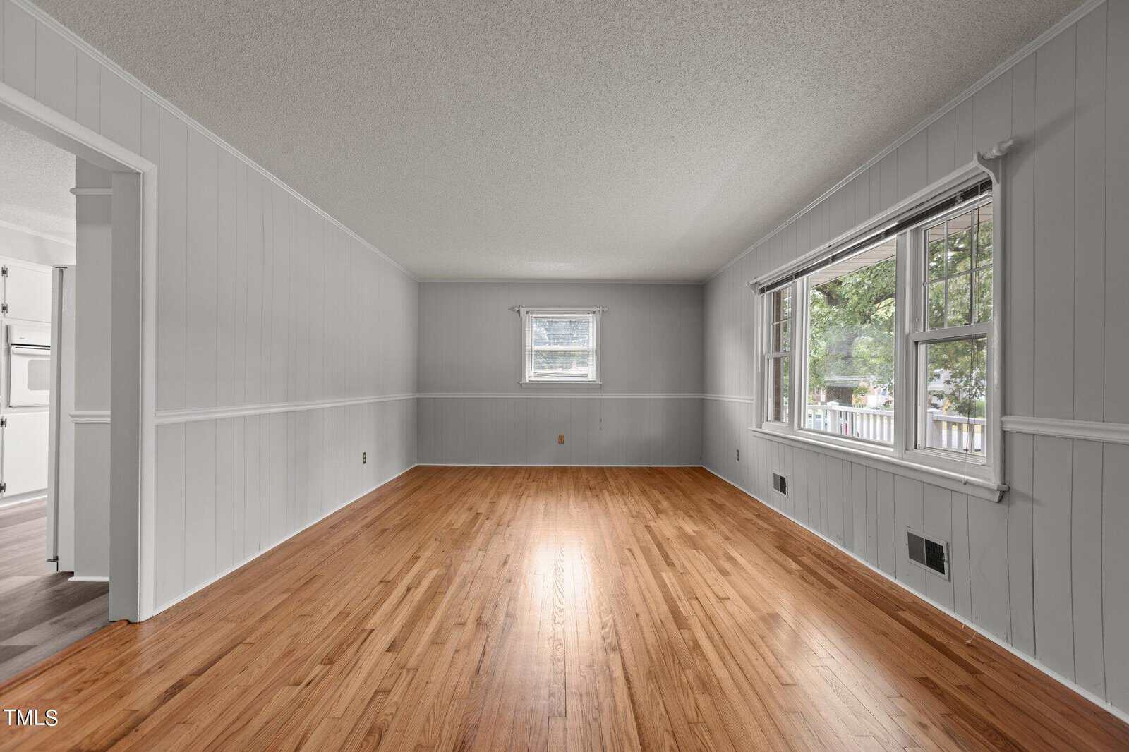 A bright, empty living room with light gray paneled walls, a large window on the right, a small window at the back, and hardwood floors. The room has a view into an adjacent kitchen area.