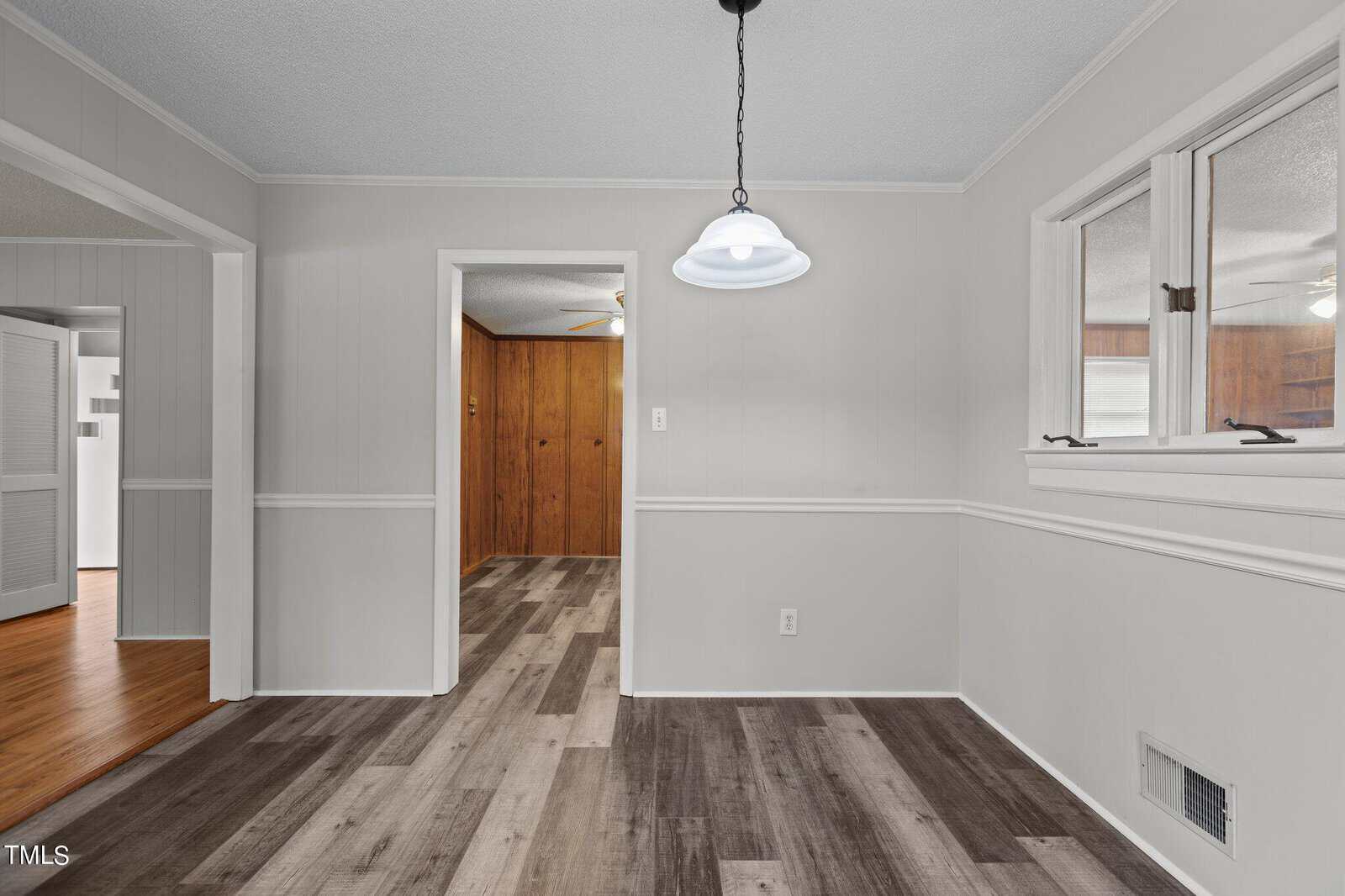 A dining area with light gray walls, a two-tone wood floor, a white pendant light, and two interior windows. Doorways lead to a wood-paneled room and a hallway with hardwood floors.