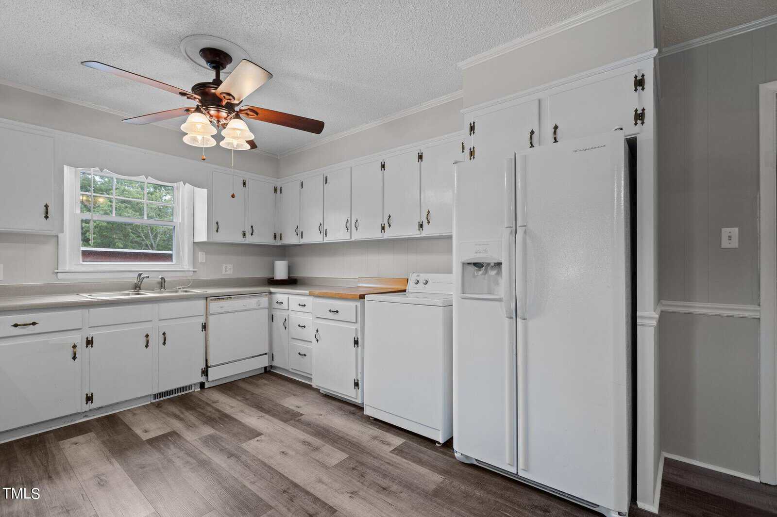 A bright, spacious kitchen with white cabinets, white appliances, a double sink under a window, light wood countertops, and wood-look flooring. A ceiling fan with lights hangs overhead.