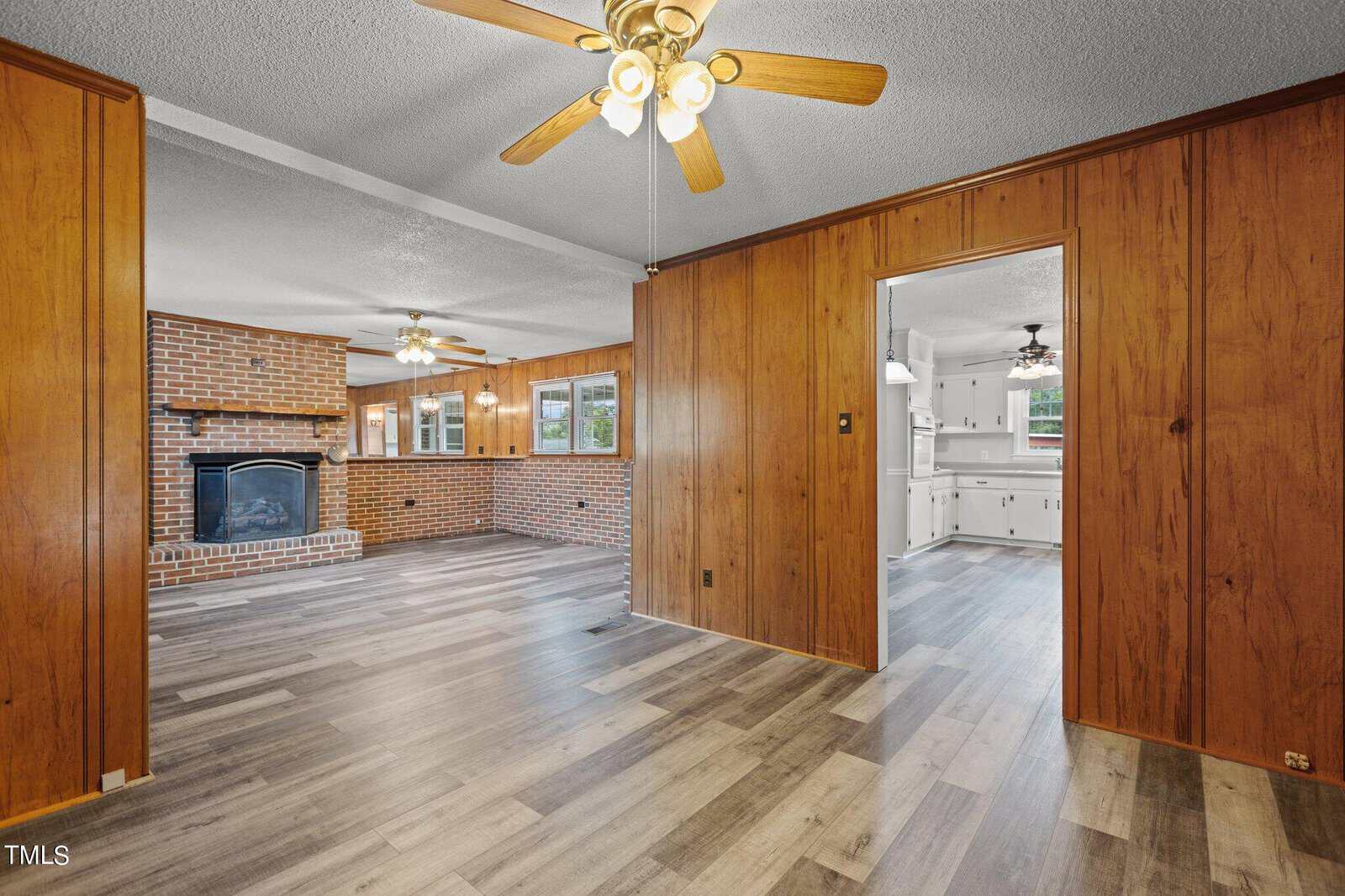 Spacious room with wood-paneled walls, two ceiling fans, large windows, and vinyl plank flooring. A brick fireplace is visible in the living area, and an adjacent kitchen can be seen through an open doorway.