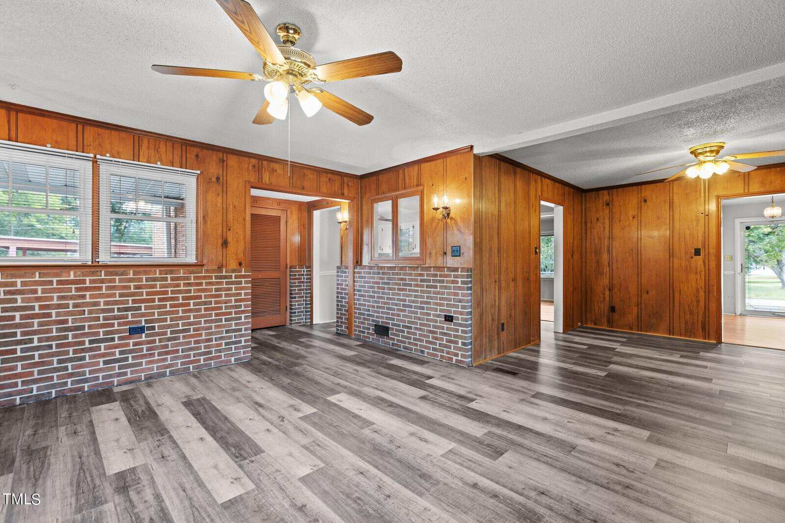 A room with wood-paneled walls, brick accents, gray wood-look flooring, ceiling fans, and large windows letting in natural light.