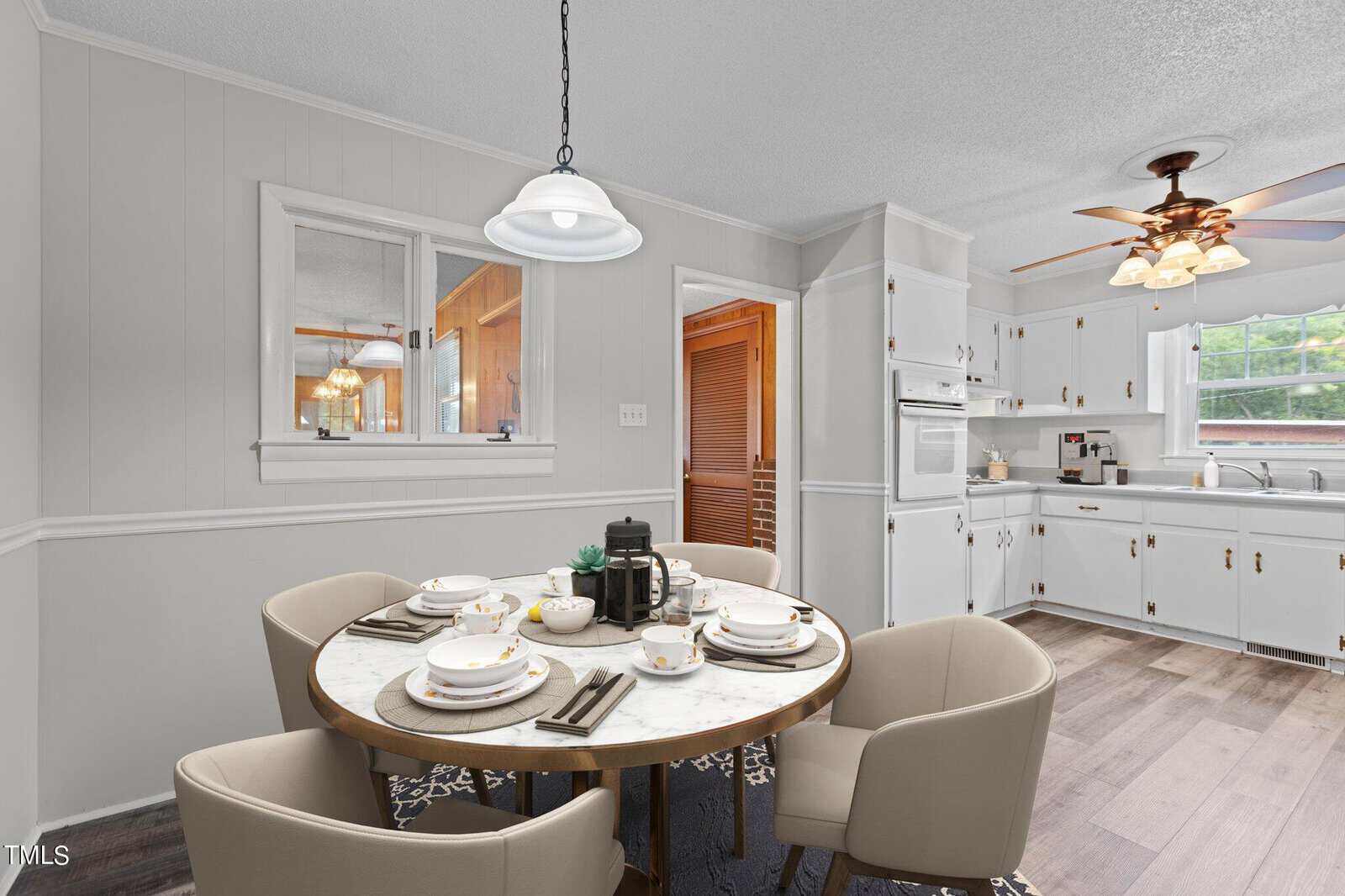 A kitchen with white cabinets and appliances, a round marble dining table set for four with beige chairs, wood-look flooring, and two hanging light fixtures. A large window and a doorway are visible in the background.