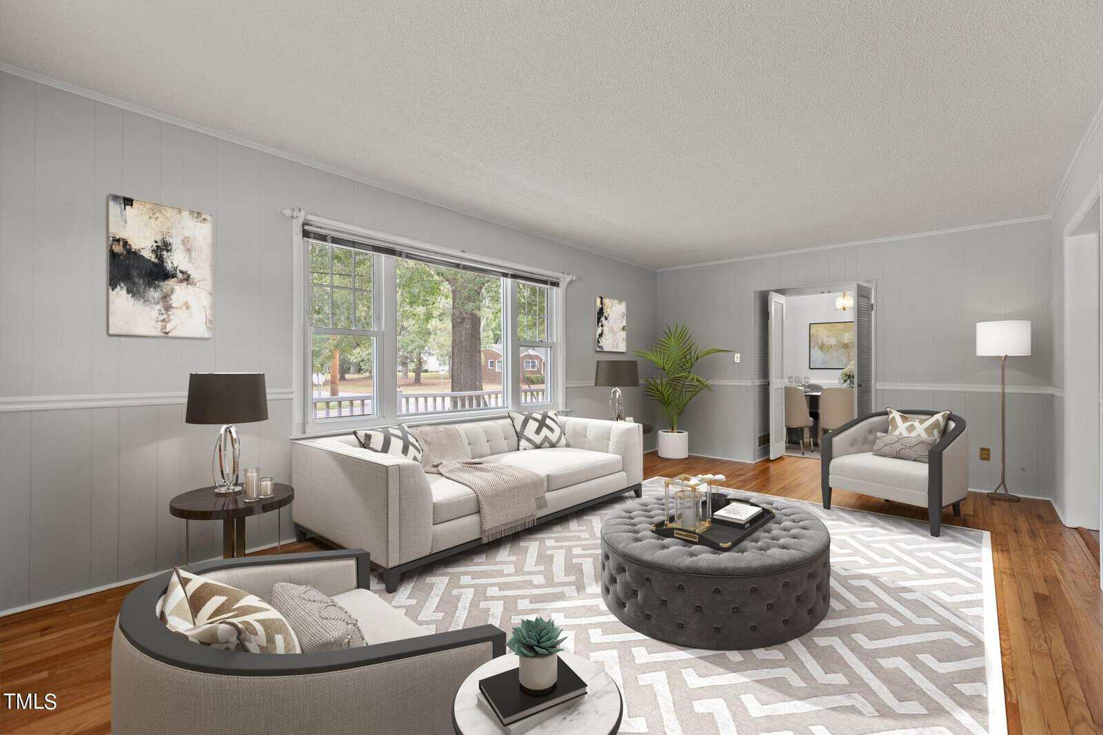 A bright living room with gray walls, hardwood floors, and large windows. The space is furnished with light-colored sofas, armchairs, a round ottoman, and abstract artwork. A plant and floor lamp add decor.