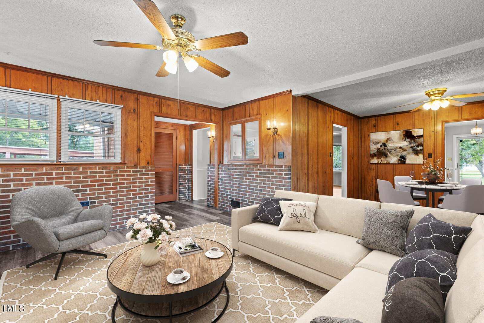 A cozy living room with wood-paneled walls, a beige sofa, a round gray chair, a patterned rug, and coffee table. Adjacent dining area, large windows, and ceiling fans provide a bright, inviting atmosphere.