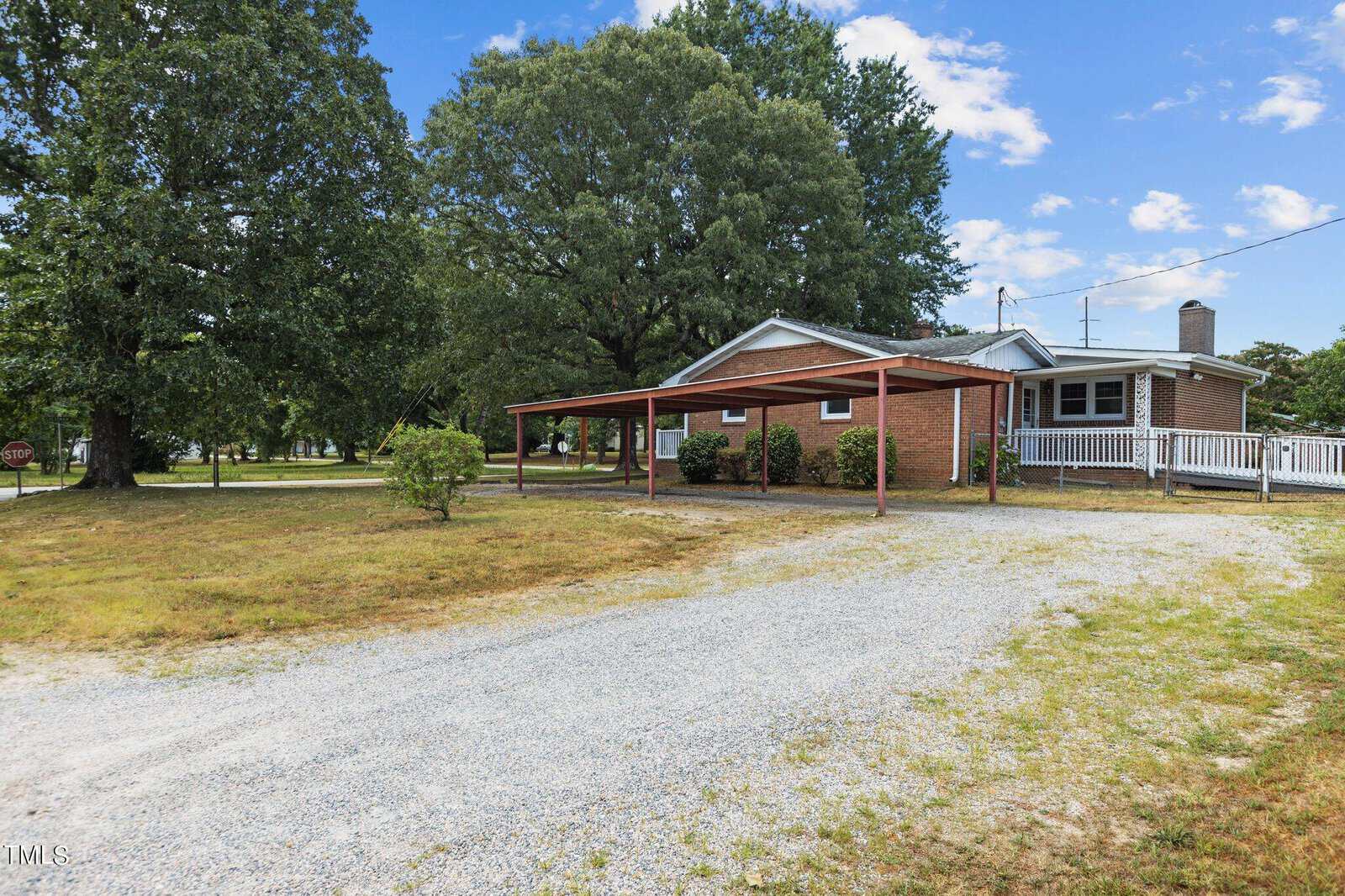 A single-story brick house with a covered carport, gravel driveway, and a large yard with trees and bushes on a sunny day.