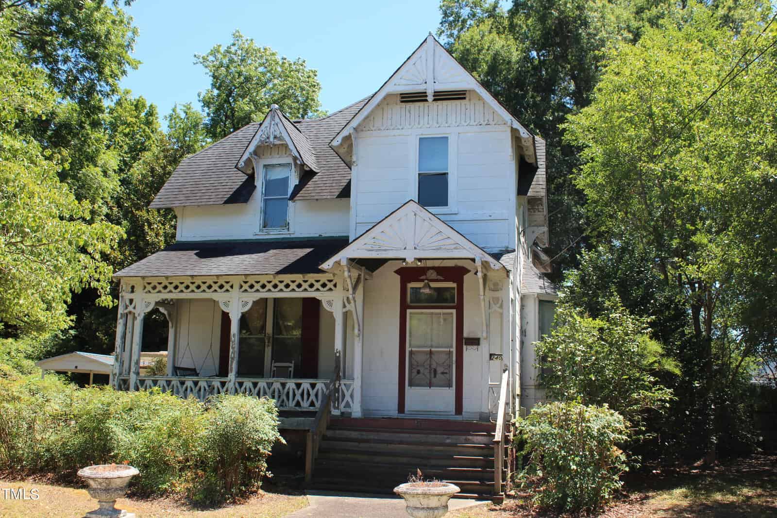 A white, two-story Victorian-style house at 240 Chavasse Avenue boasts a steeply pitched roof, detailed woodwork on the front porch, and an arched window. Surrounded by trees and bushes in Henderson, NC, it features a concrete pathway leading to the front steps.
