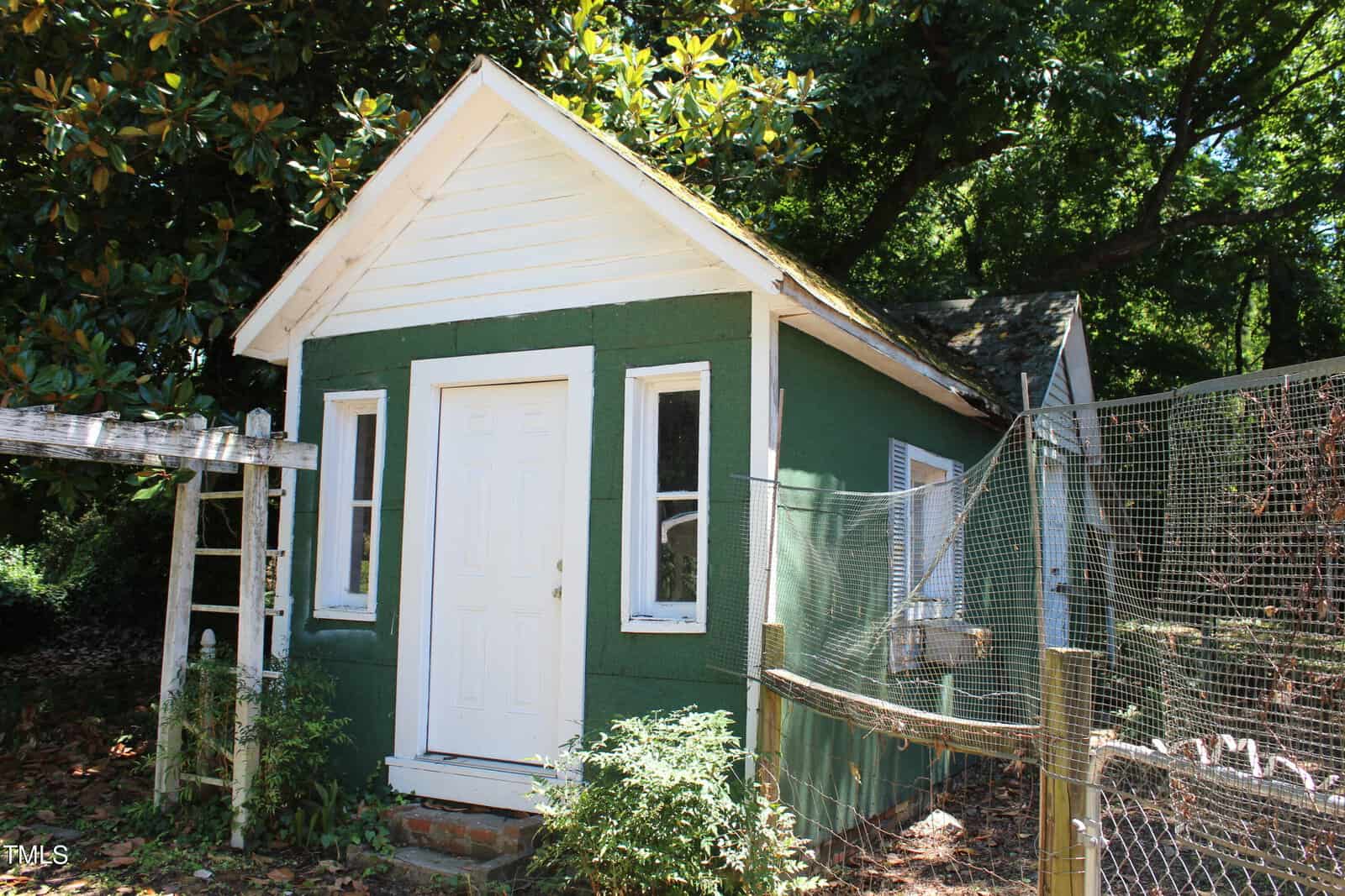 A small green shed with a white door and window frames stands in a garden area. A white wooden trellis is attached beside it, and a wire mesh fence encloses a section on the right. The shed is shaded by large trees, and sunlight filters through the foliage.