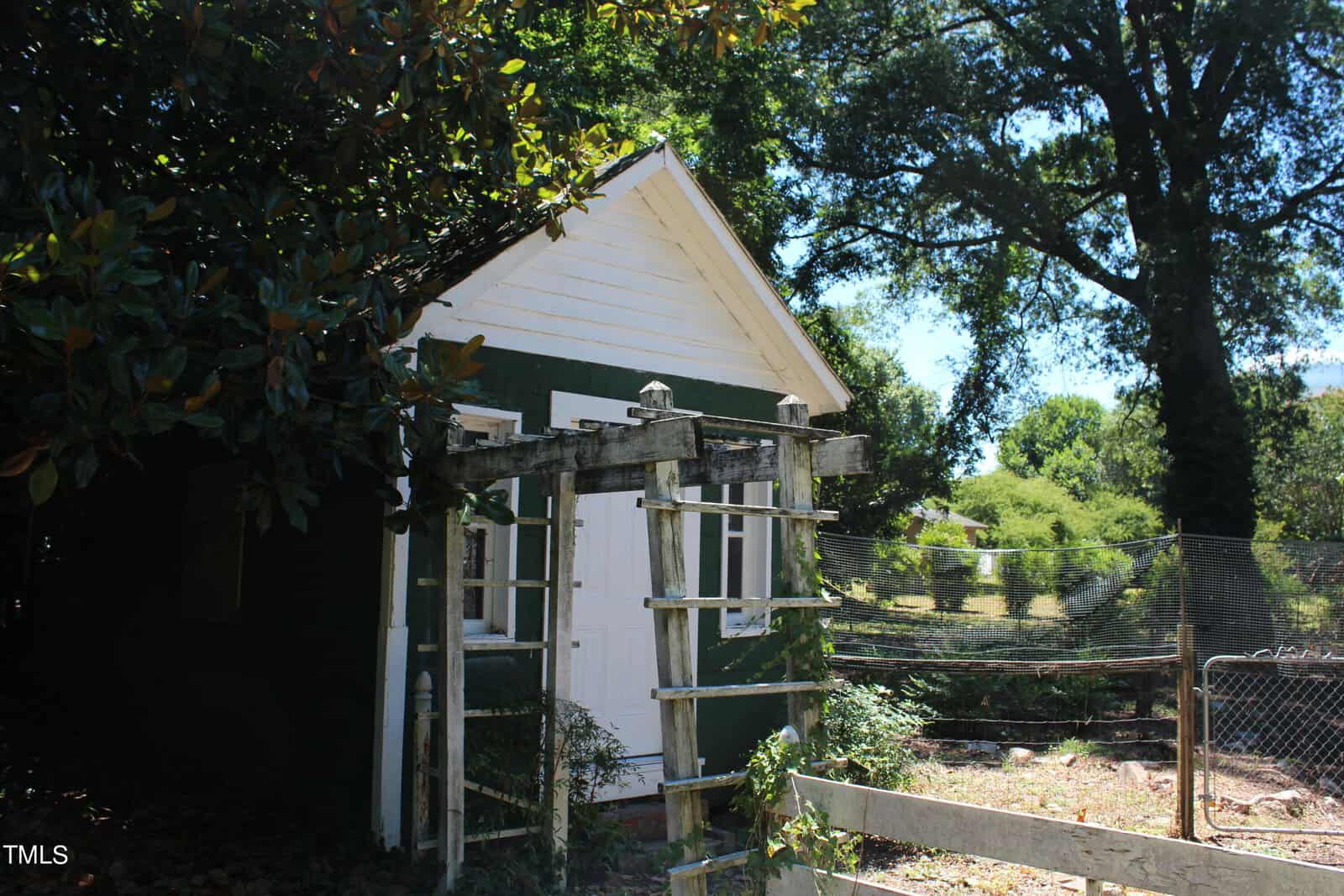 A small white and green shed with a peaked roof stands in a garden. In front of the shed is a weathered wooden trellis. The area is surrounded by lush trees and greenery, with a wire fence visible on the right side of the image.