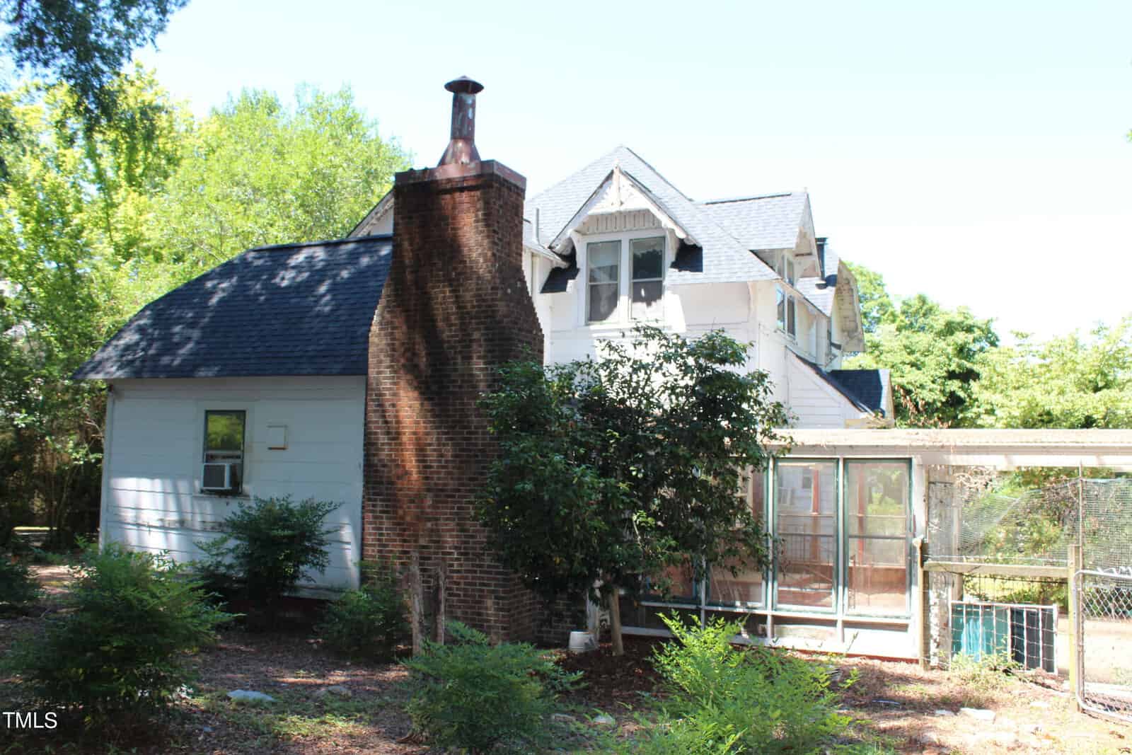 A two-story house with white siding, dark shingles, and a prominent brick chimney on its side. The house features a screened-in porch, surrounded by lush green trees and bushes, with a clear blue sky overhead.