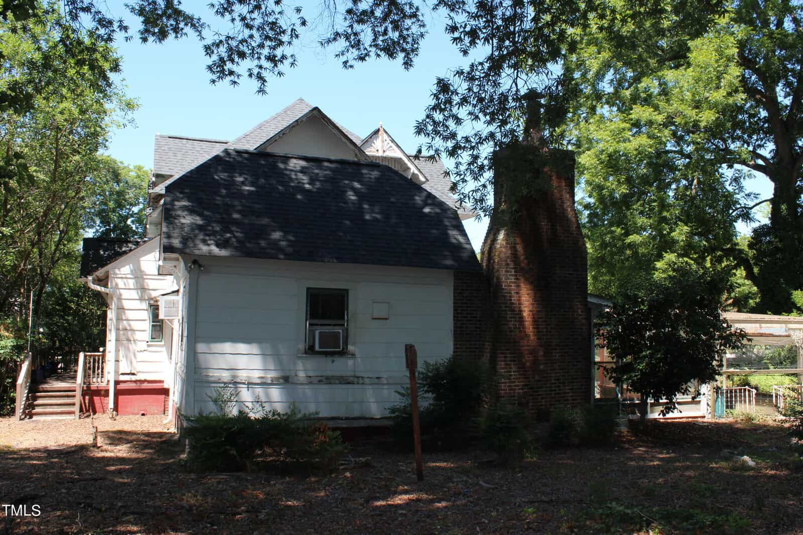 A two-story house with white siding and a shingled roof. There is a prominent brick chimney on the right side. The house is surrounded by trees, casting shadows on the scene. An air conditioning unit is visible in a window. The yard has wooden steps leading up to a deck.