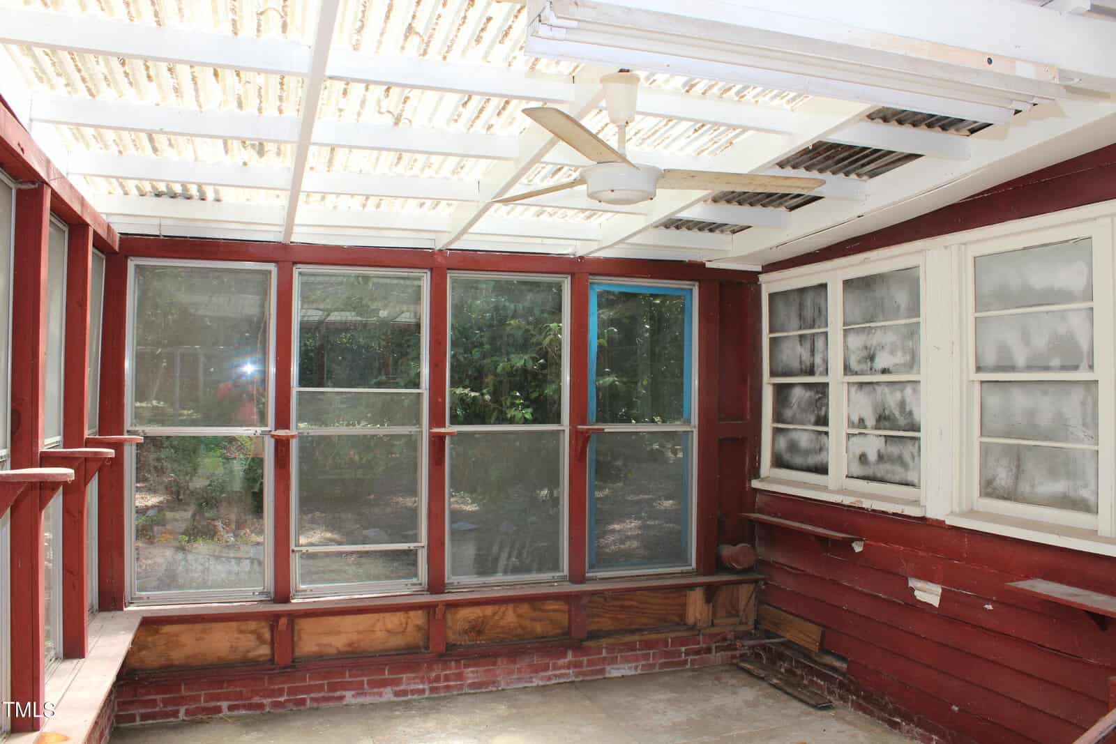 A sunroom with red wooden walls features multiple large windows, some screened and some glass, allowing sunlight to stream in. The ceiling has a white pergola style with clear panels and includes a ceiling fan. The floor is bare concrete, and a garden is visible outside.