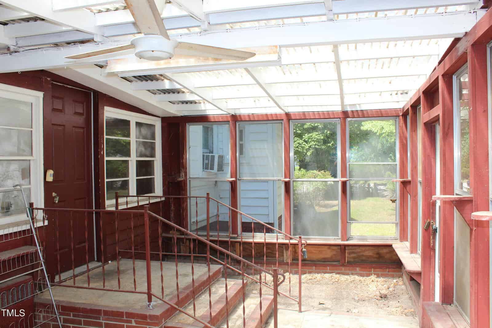 A small enclosed porch with red walls and a transparent roof. The porch features glass windows, a door, and an overhead ceiling fan. There are metal railings and steps leading down to a concrete floor. Sunlight streams through the roof, illuminating the space.