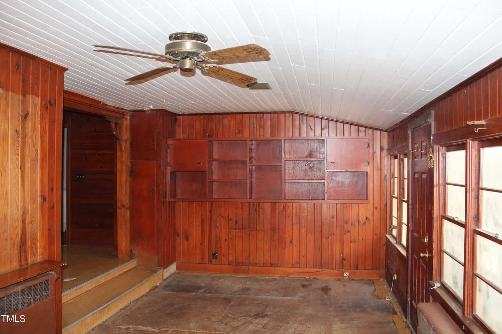 A rustic wood-paneled room with empty shelving units on one wall, a ceiling fan, and large windows. The floor appears unfinished, and there is a small step leading to another room. The ceiling is white, contrasting with the wooden walls and trim.