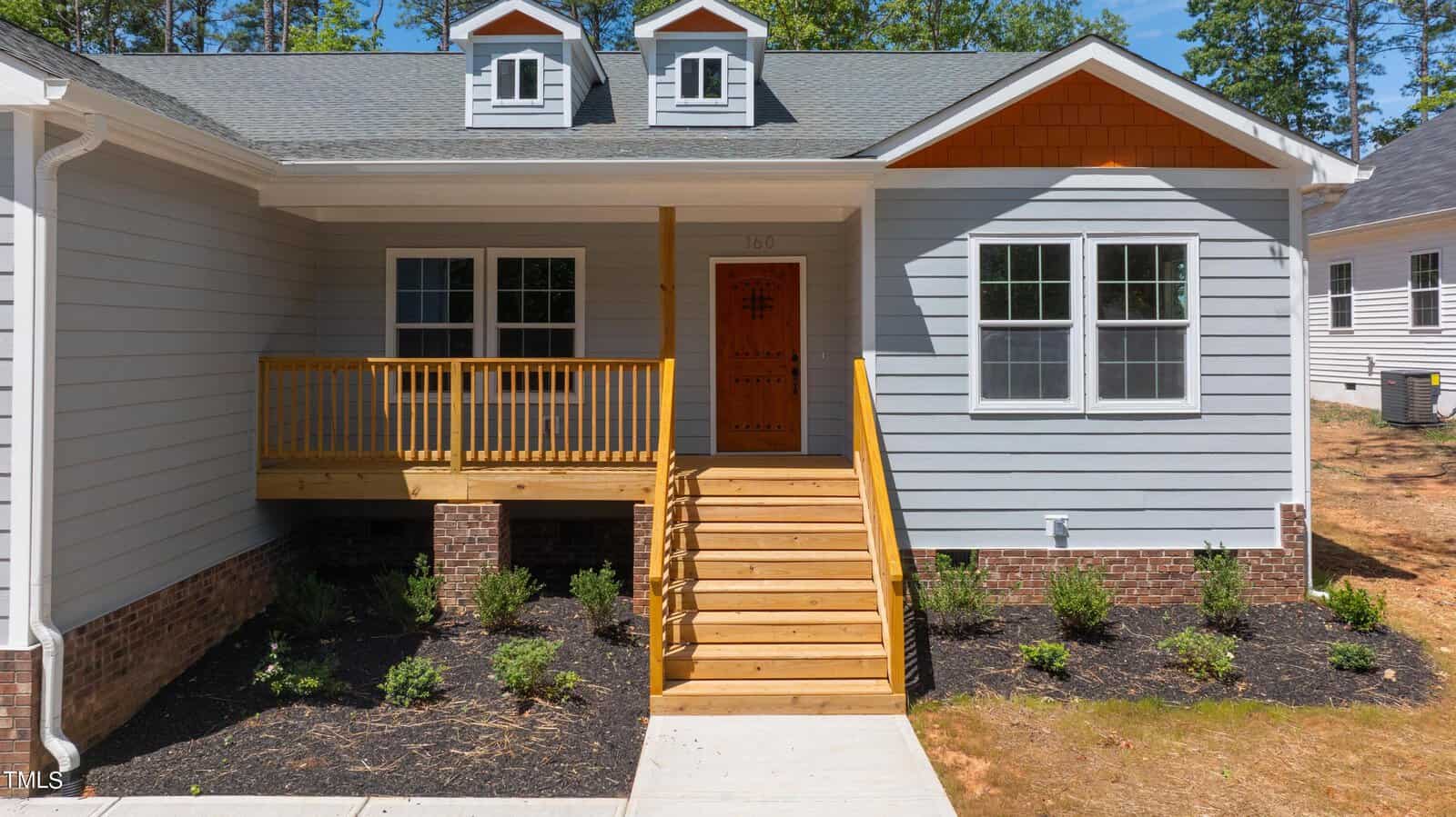 A modern gray house with white trim, a front porch with wooden railings and steps, a wooden front door, brick foundation, and neatly landscaped shrubs, under a clear sky.