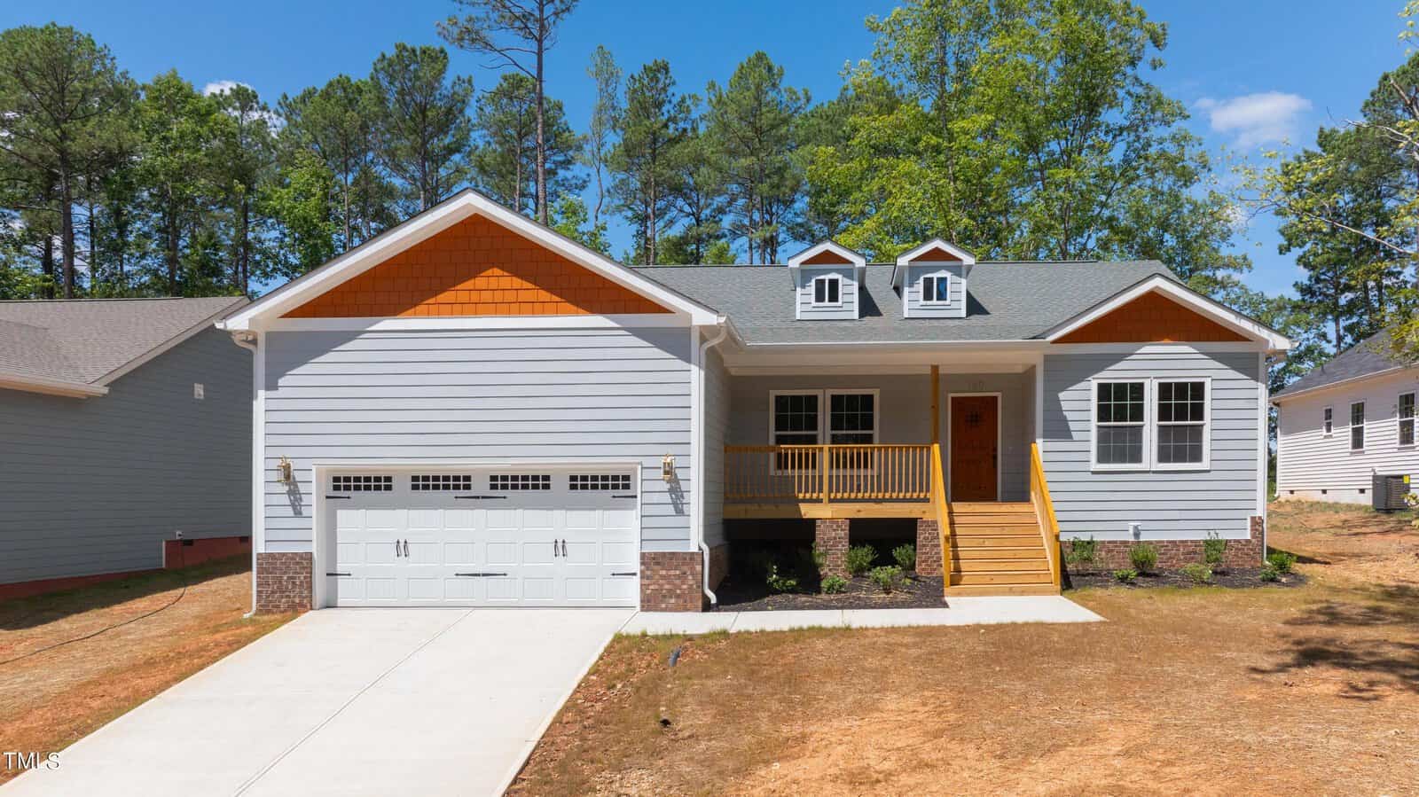 A modern single-story house with gray siding, orange gable accents, a two-car garage, a front porch with wooden steps and railing, and a concrete driveway, surrounded by trees and neighboring homes.