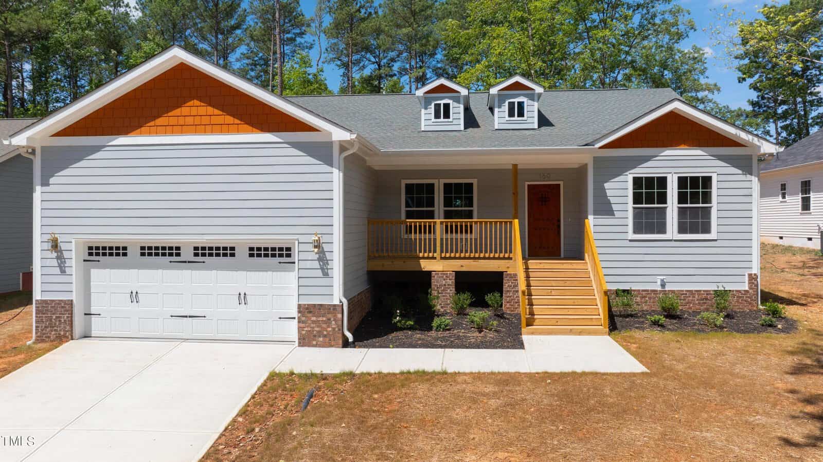 Single-story house with light gray siding, a front porch with wooden steps and railing, double garage doors, and two dormer windows on the roof, surrounded by trees and a partially landscaped yard.