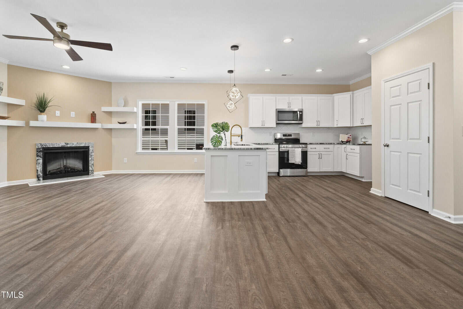 Modern, open-concept kitchen and living area with light walls, wood flooring, white cabinets, stainless steel appliances, a central island, built-in shelves by a fireplace, and large windows with shutters.