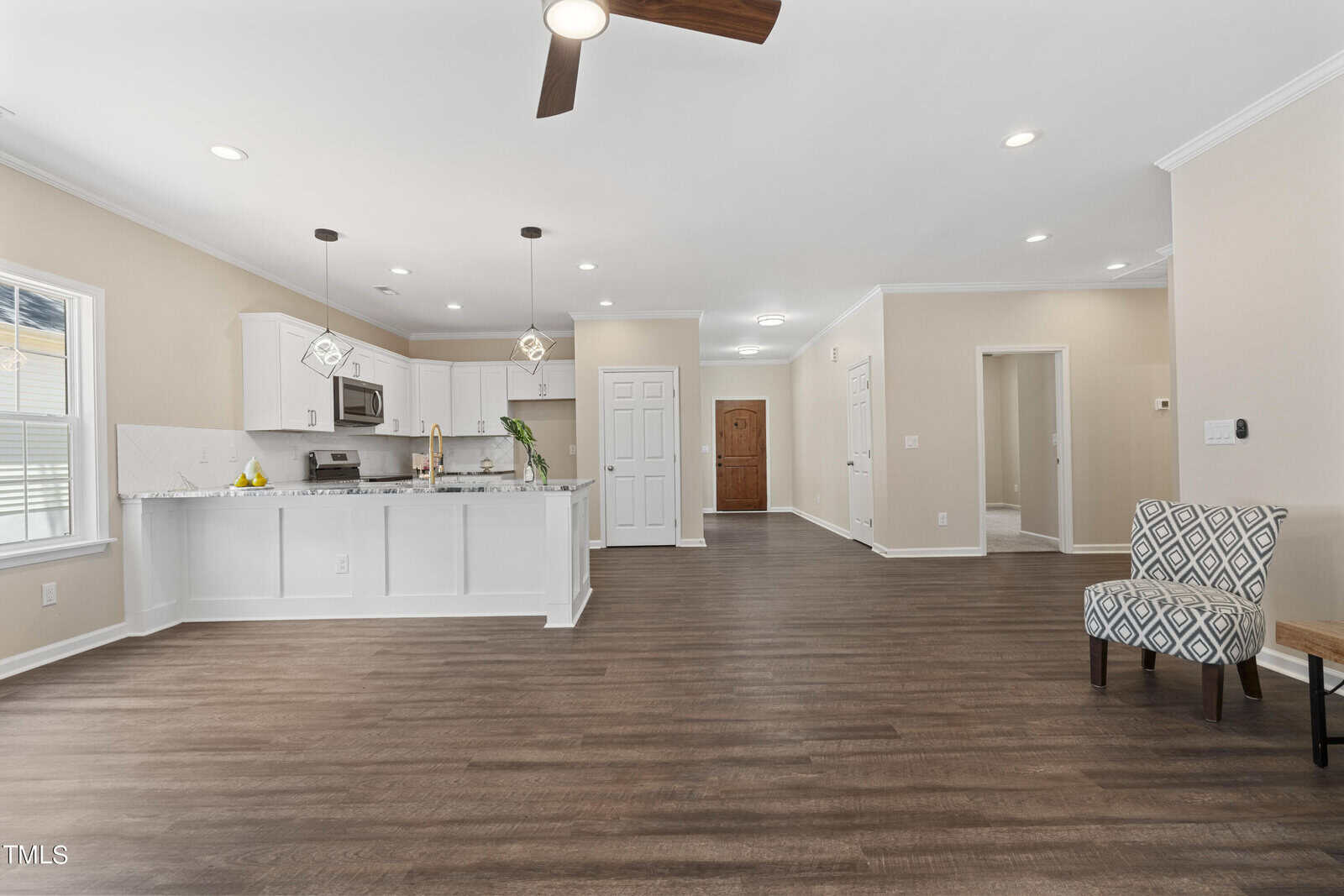 Open-concept living space with wood flooring, white kitchen cabinets, stainless steel appliances, central ceiling fan, patterned armchair, and neutral walls, featuring ample natural light from multiple windows.