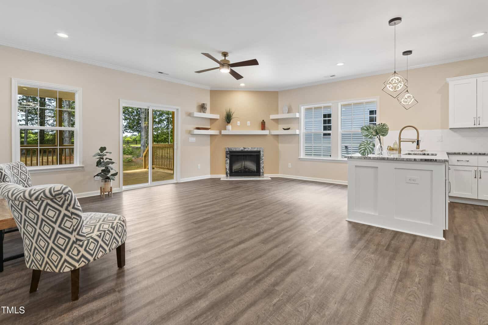 Spacious, modern living area with wood flooring, a patterned armchair, fireplace, open kitchen with white cabinets, pendant lights, large windows, and glass doors leading to a deck with trees visible outside.