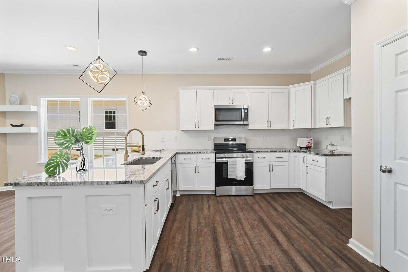 Modern kitchen with white cabinets, stainless steel appliances, granite countertops, pendant lights, a large island with a sink, wooden floors, and neutral walls. A plant in a vase decorates the island.
