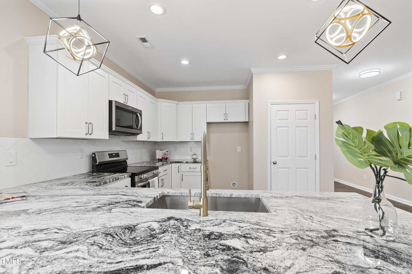 Modern kitchen with white cabinets, stainless steel appliances, a marbled gray and white countertop, gold faucet, geometric pendant lights, and a green plant in a glass vase on the island.