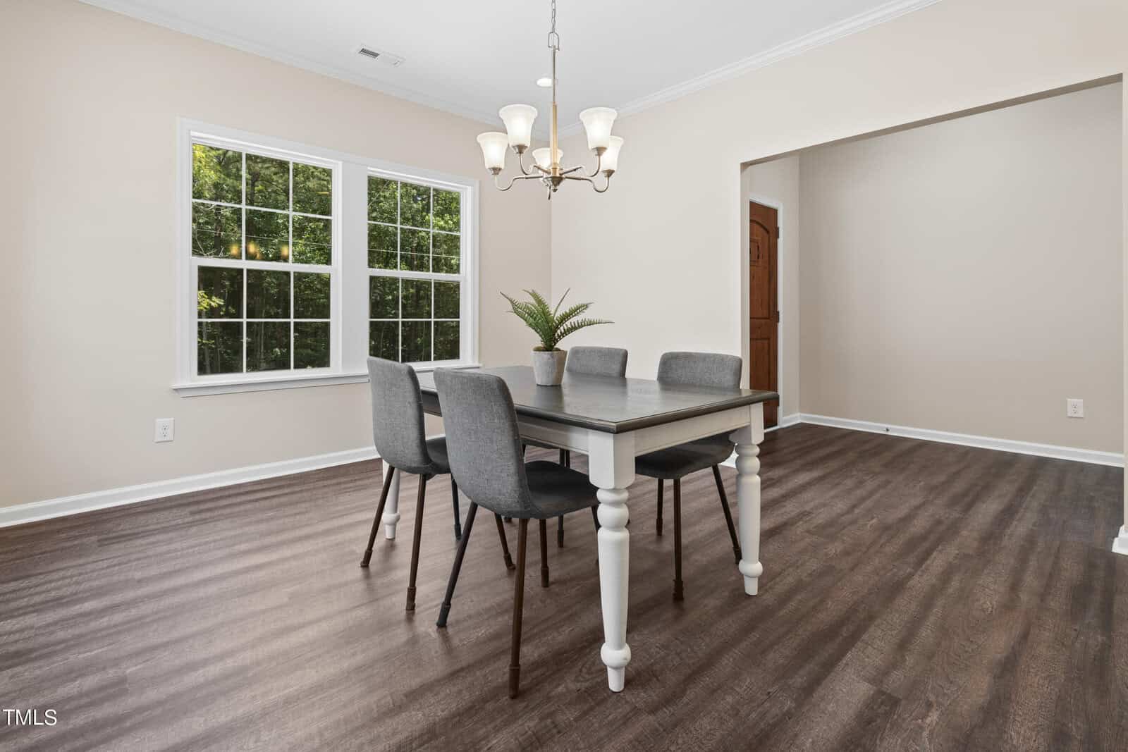 A dining room with four gray chairs around a dark table, a potted plant centerpiece, wood flooring, beige walls, large window, and chandelier overhead.