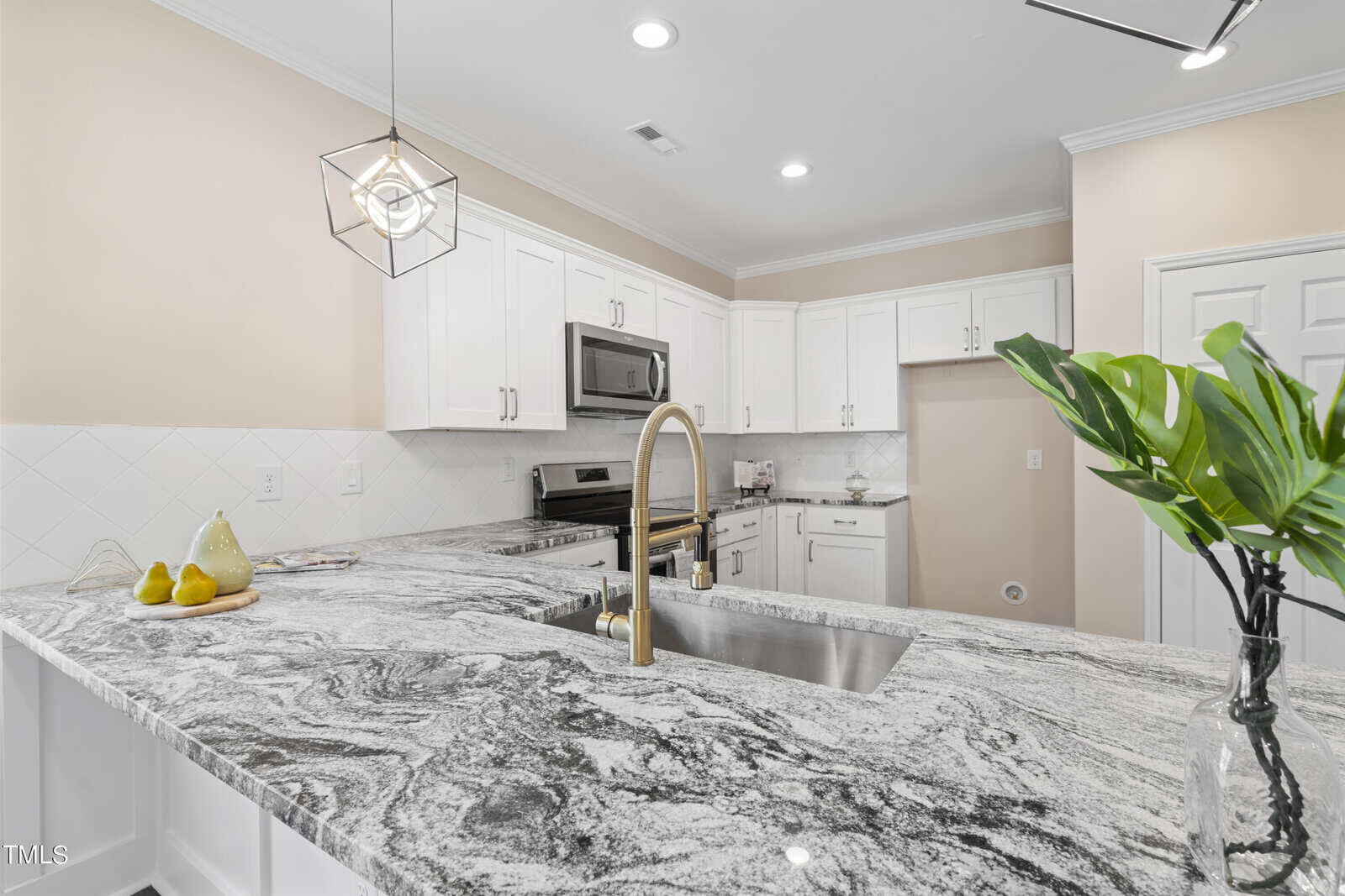 Modern kitchen with white cabinets, stainless steel appliances, a marble-patterned countertop, built-in sink with gold faucet, decorative plant, and a tray with pears and cups. Soft beige walls and recessed lighting brighten the space.