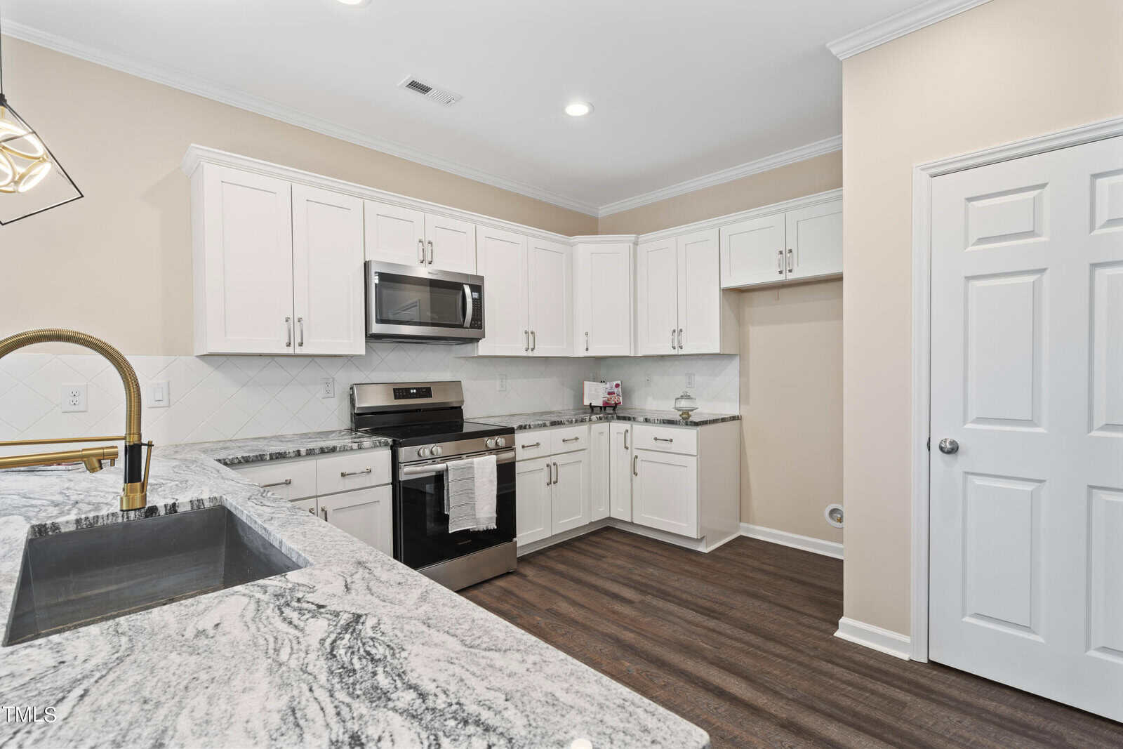 Modern kitchen with white cabinets, stainless steel appliances, marble countertops, tile backsplash, and wood flooring. There is a built-in space for a washer and dryer and a gold faucet over a large sink.