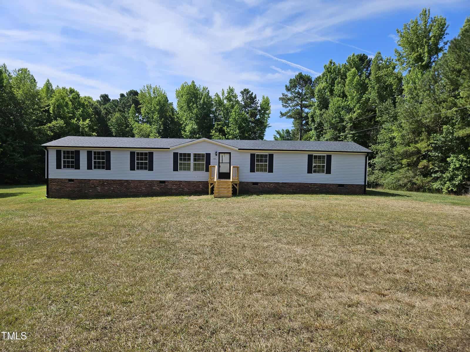 A single-story house at 2142 Mountain Creek Road features a dark roof and light-colored siding, enveloped by a grassy lawn and tall green trees. The Oxford blue sky is peppered with clouds, and a small wooden porch with steps leads to the inviting entrance.