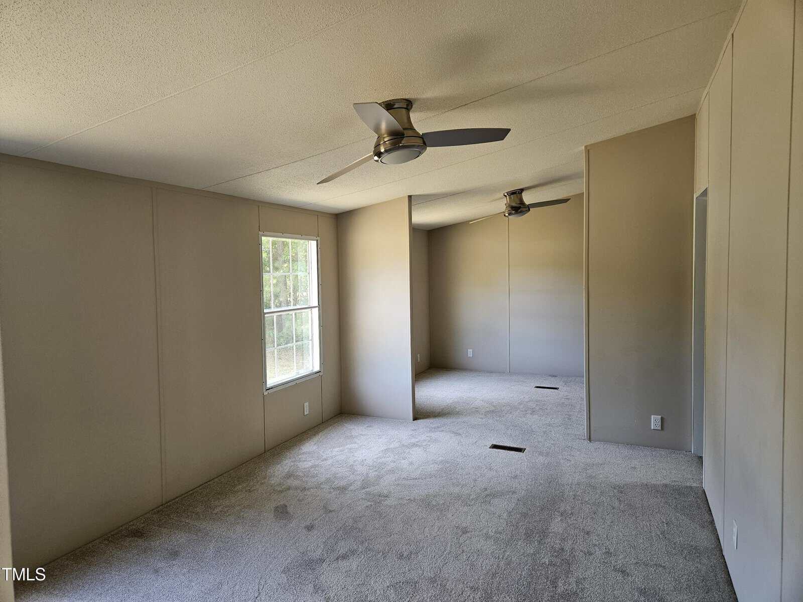 A spacious, empty room with beige walls and carpeted flooring. Two ceiling fans are installed, and sunlight streams through a window on the left, illuminating the space.