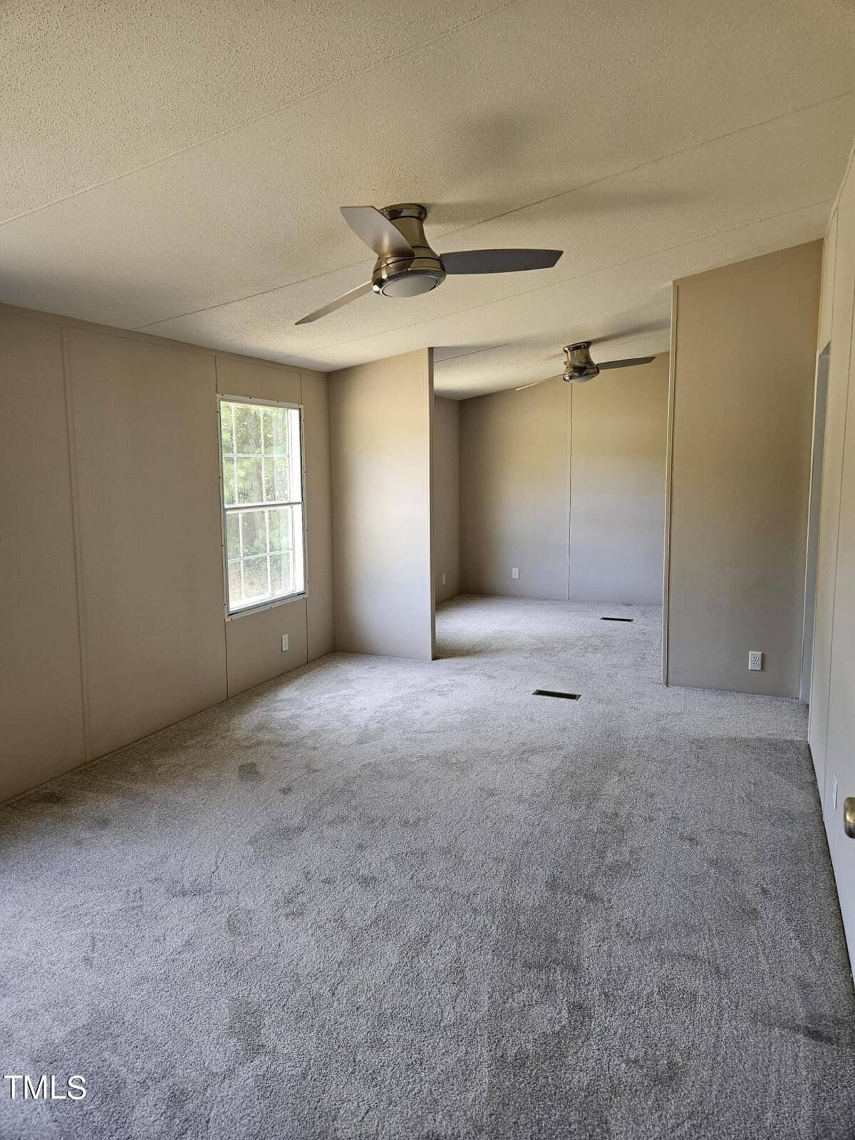 An empty room with beige walls and carpeting, featuring two ceiling fans and large windows letting in natural light. The room is long and narrow, with modern fixtures and minimal decor.