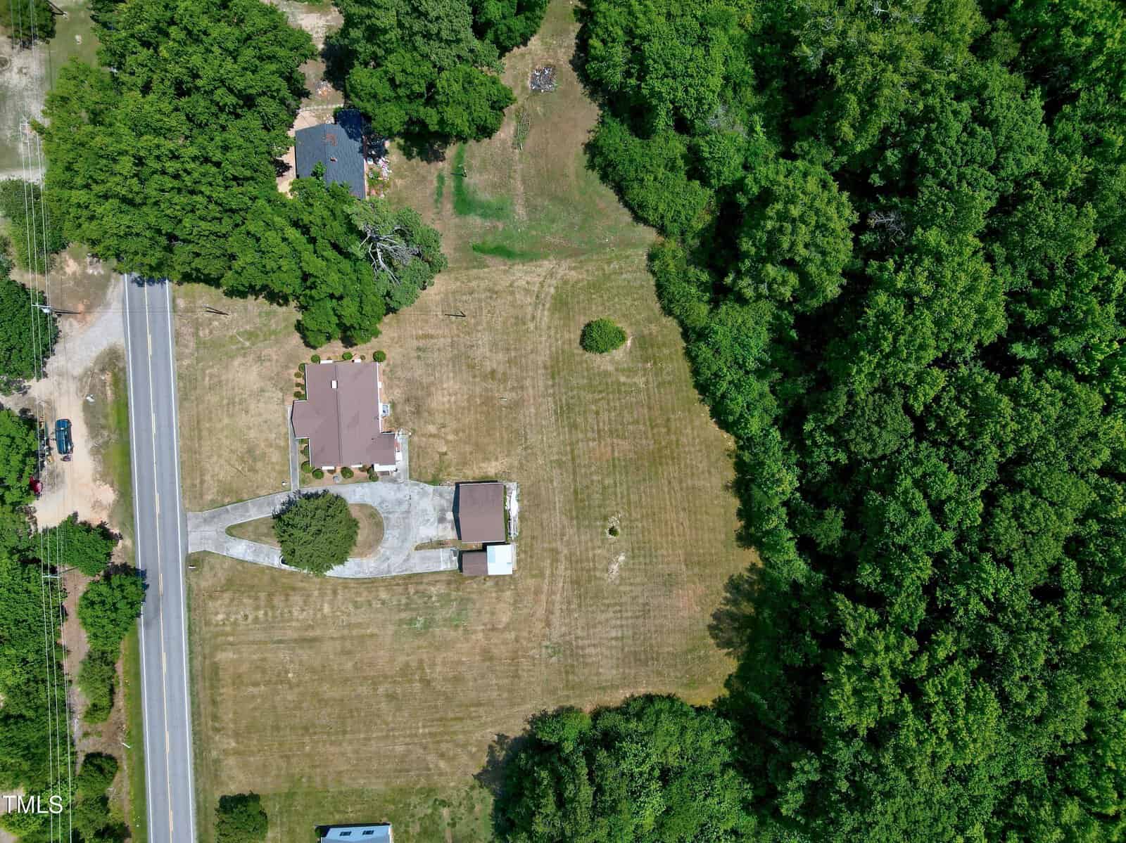 An aerial view of a property featuring a house with a brown roof, a driveway leading to the road, and a large grassy yard bordered by dense green trees. The house is surrounded by greenery, and a smaller structure is visible near the house.