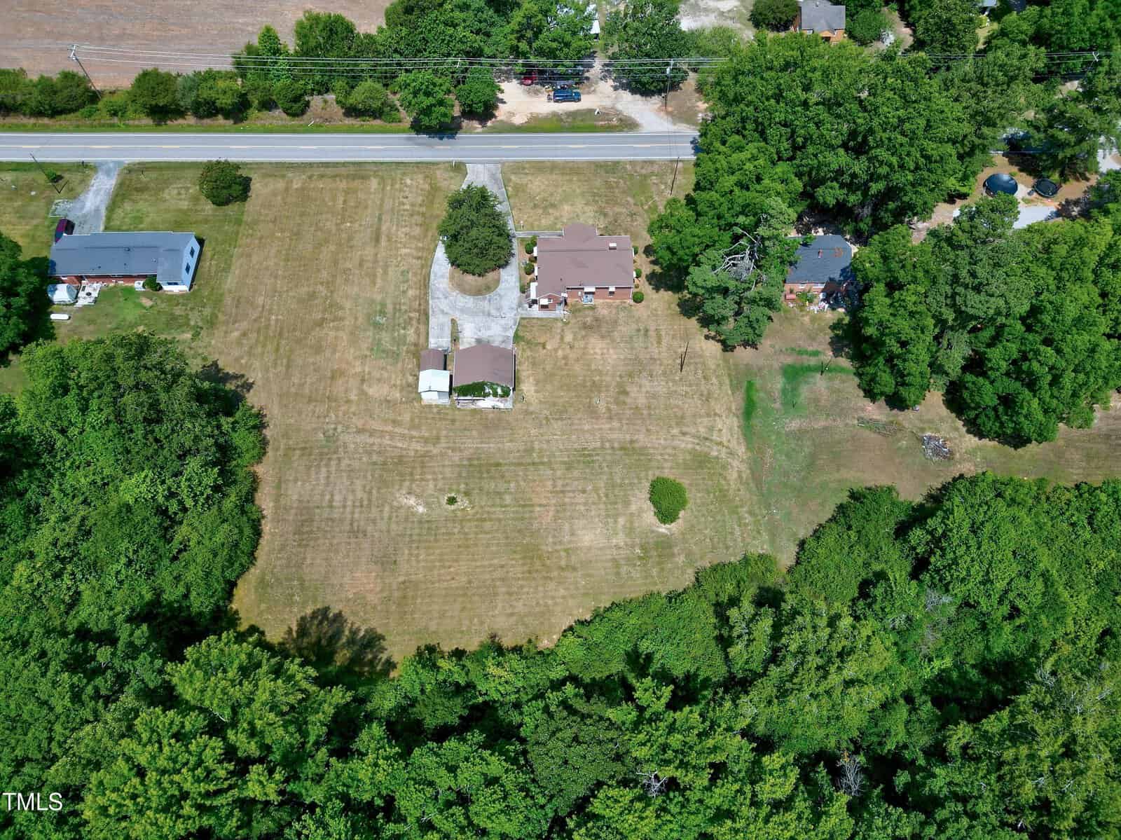 An aerial view of a rural property shows a large, clear section of land with scattered trees. The property includes a single-story house, an outbuilding, and a gravel driveway. It is bordered by a road at the top and surrounded by lush green trees and other properties.