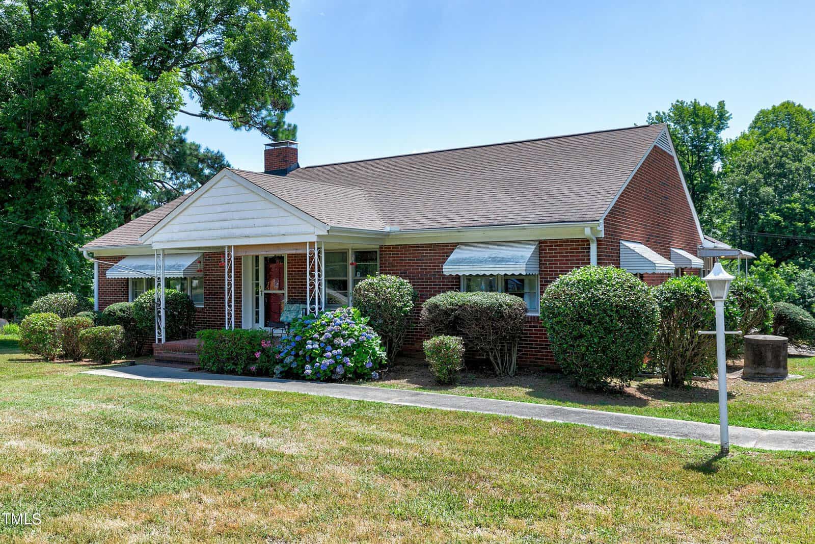 A single-story red brick house with a triangular white porch roof supported by decorative metal posts. The home is surrounded by neatly trimmed bushes and a well-maintained lawn. A white lamp post is positioned near the walkway leading to the front entrance.