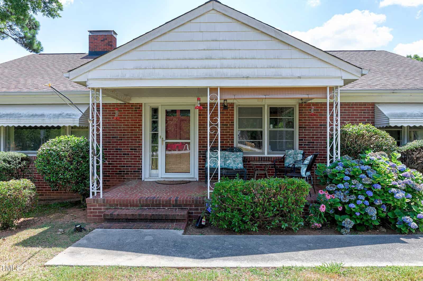 A one-story brick house with a front porch supported by decorative metal columns. The porch is furnished with a wicker loveseat, a chair, and a small table. Bushes and blooming hydrangeas surround the walkway leading to the front door. The sky is clear and sunny.