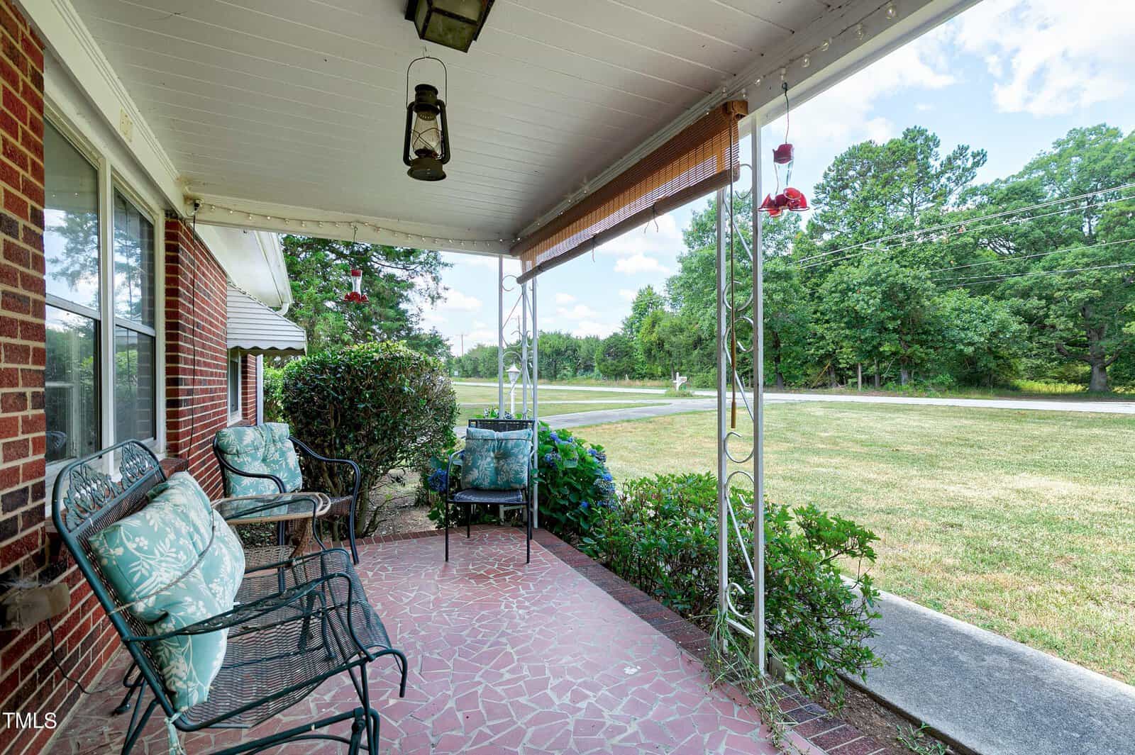 A cozy porch with cushioned chairs and a small bench, framed by a white roof and supported by decorative iron columns. Red brick walls complement the stone-tiled floor. The porch overlooks a grassy yard with trees and a street visible in the distance under a blue sky.
