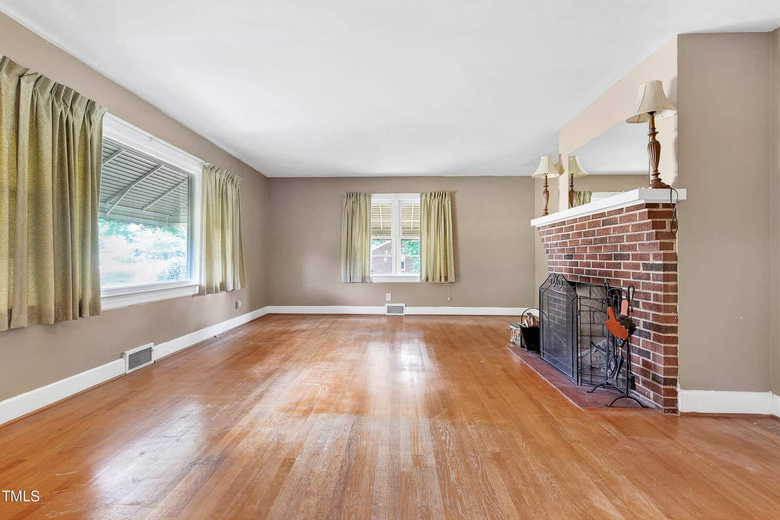 A spacious living room with hardwood floors, beige walls, and large windows with yellow curtains. There is a red brick fireplace with a black metal guard on the right, topped with a mirror and lamps. Natural light fills the room, creating a warm atmosphere.