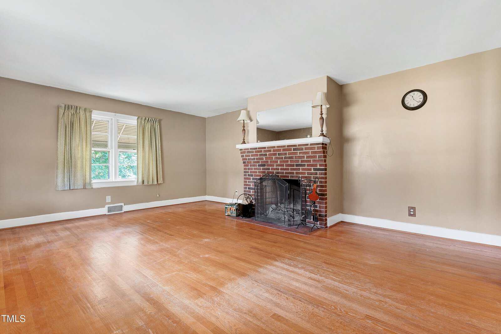 Spacious living room with hardwood floors, a brick fireplace, two windows with beige curtains, a wall clock, and neutral-colored walls.