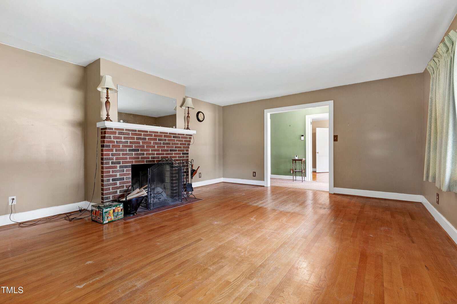 A spacious living room with hardwood floors, a brick fireplace with a mirror above, two lamps, beige walls, and an open doorway leading to a green-painted room.