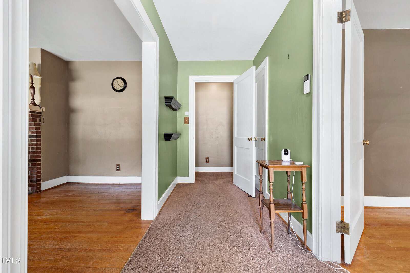 A hallway with green walls and tan carpet, leading to open doors. A small wooden table with a security camera sits by the wall. Adjacent room with wood flooring, fireplace, and a wall clock are visible.