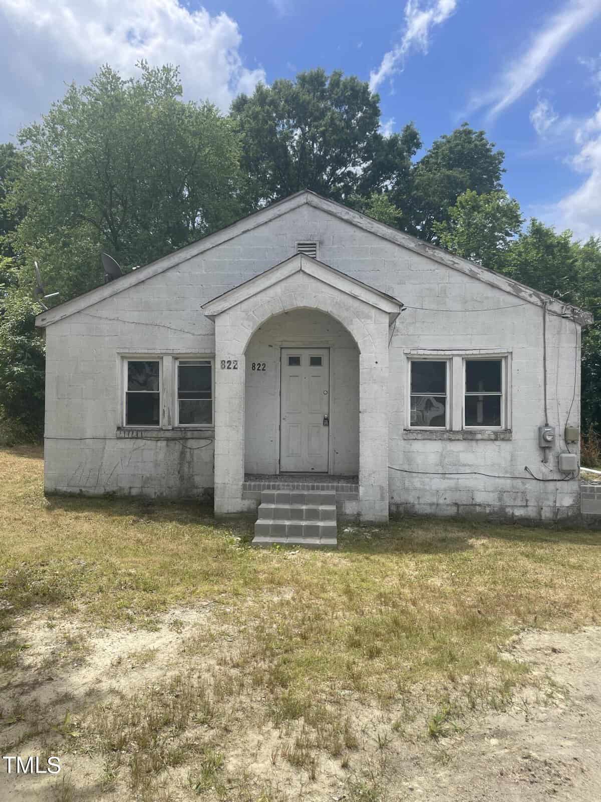 A weathered, one-story white house at 822 Eaton Street in Henderson, NC, features a central arched entrance and a small set of steps leading up to it. With two windows on either side of the doorway, it is surrounded by grass and a few tall trees under a partly cloudy sky.
