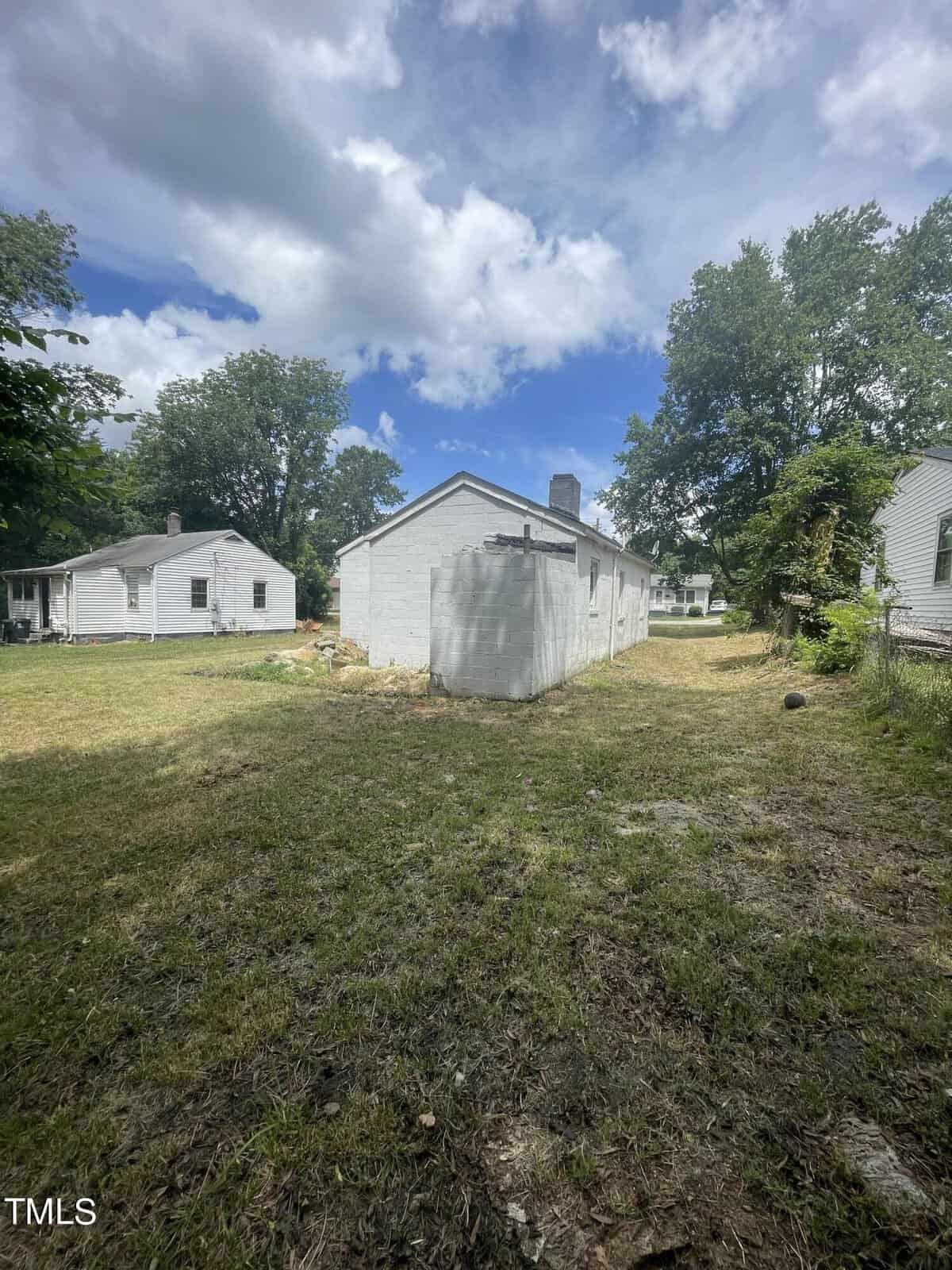 A backyard view featuring a weathered, white-painted shed with a sloped roof in the foreground. The yard is mostly grass with patches of dirt. Trees and other white houses are visible in the background under a partly cloudy sky.
