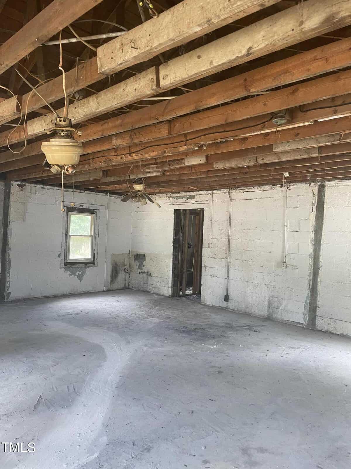 An unfinished room with exposed wooden beams on the ceiling and a concrete floor. There are white, unpainted brick walls with a single window on the left side and a doorway in the middle. The room appears to be under renovation or construction.