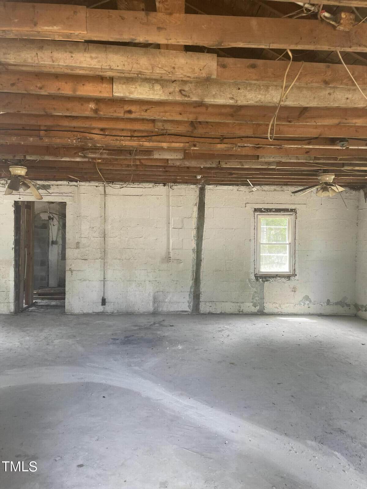 An empty, unfinished room with a concrete floor, exposed wooden ceiling beams, and a brick wall with visible wear and cracks. There's one window on the right, an open doorway on the left, and ceiling fans without blades hanging from the ceiling.