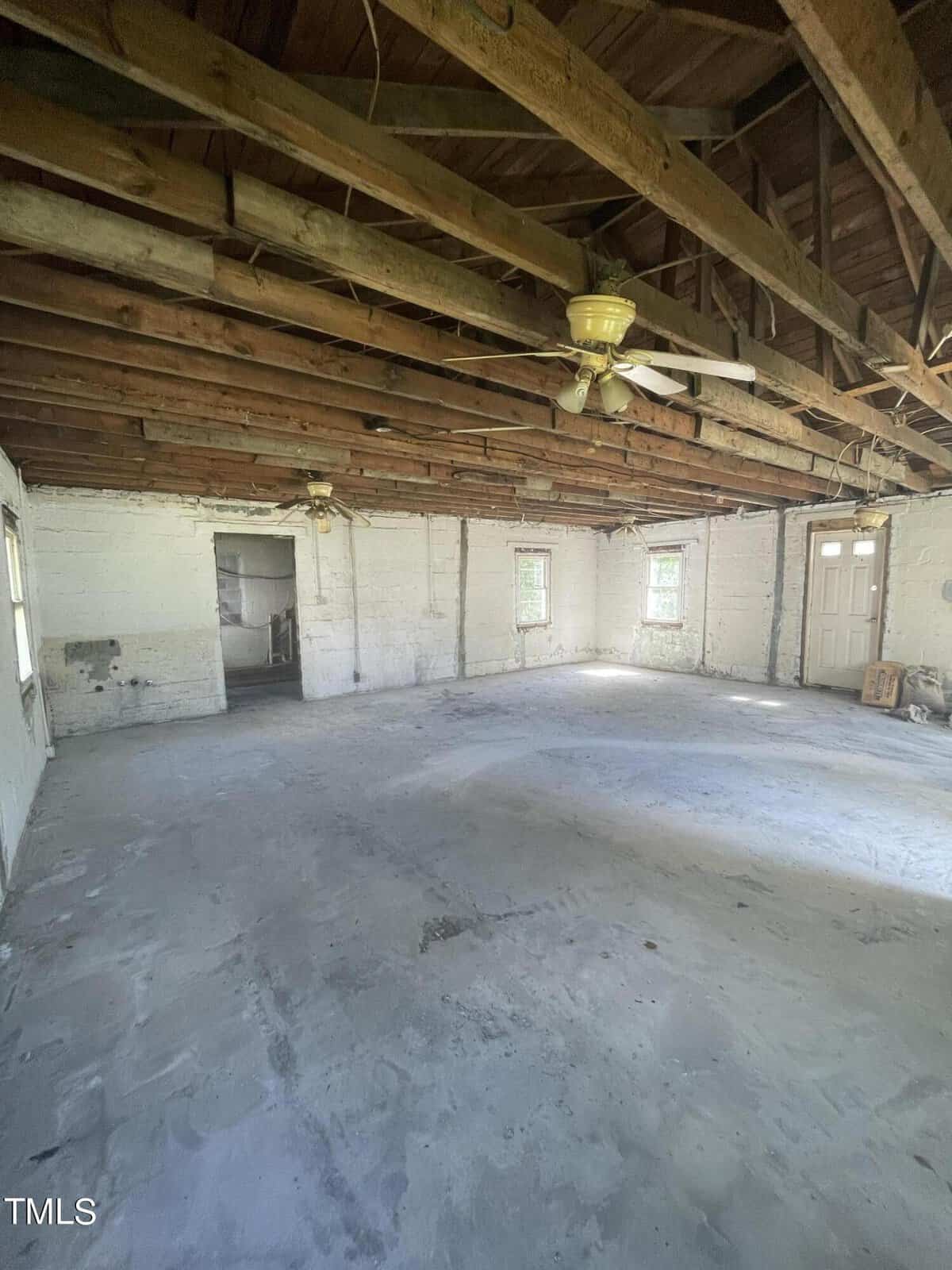 Image of a large, unfinished room with visible wooden ceiling beams and exposed brick walls. The concrete floor appears slightly uneven and remnants of construction materials are scattered. There are multiple small windows and a ceiling fan in the center.