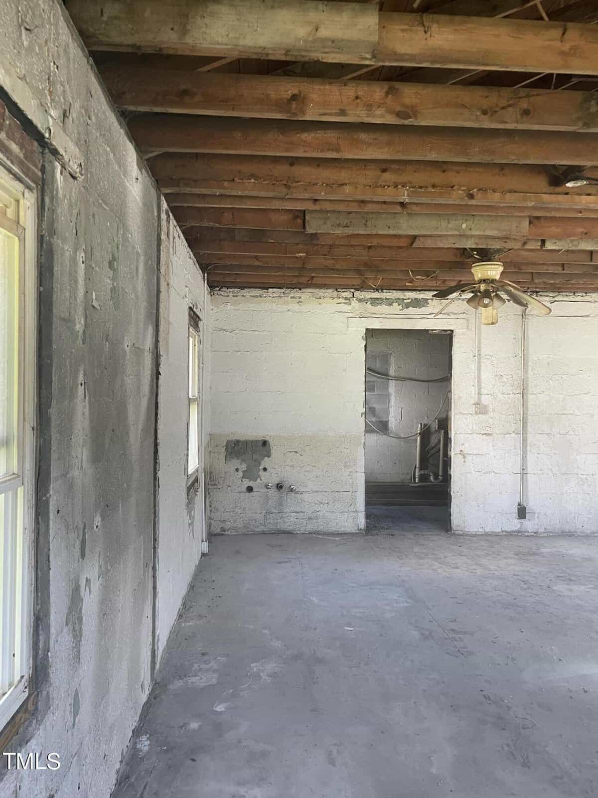 A bare room with exposed wooden ceiling beams and unpainted concrete walls. The floor is unfinished concrete, and there is a ceiling fan. A doorway leads to another room, and there are two windows on the left wall.